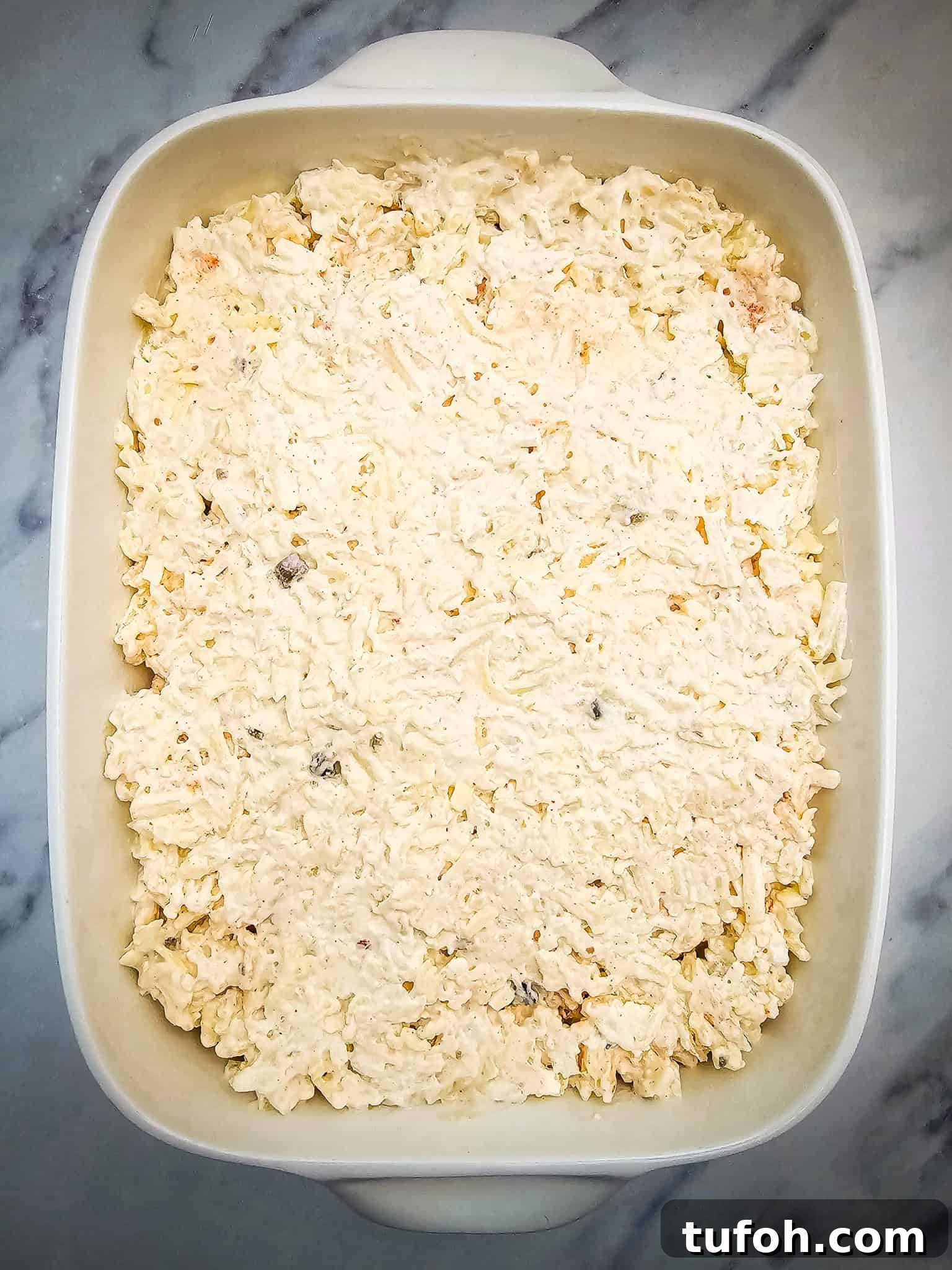 Creamy Cheesy Potato Casserole 6 A white casserole dish filled with an uncooked creamy cheese and hashbrown mixture sits on a marble surface.