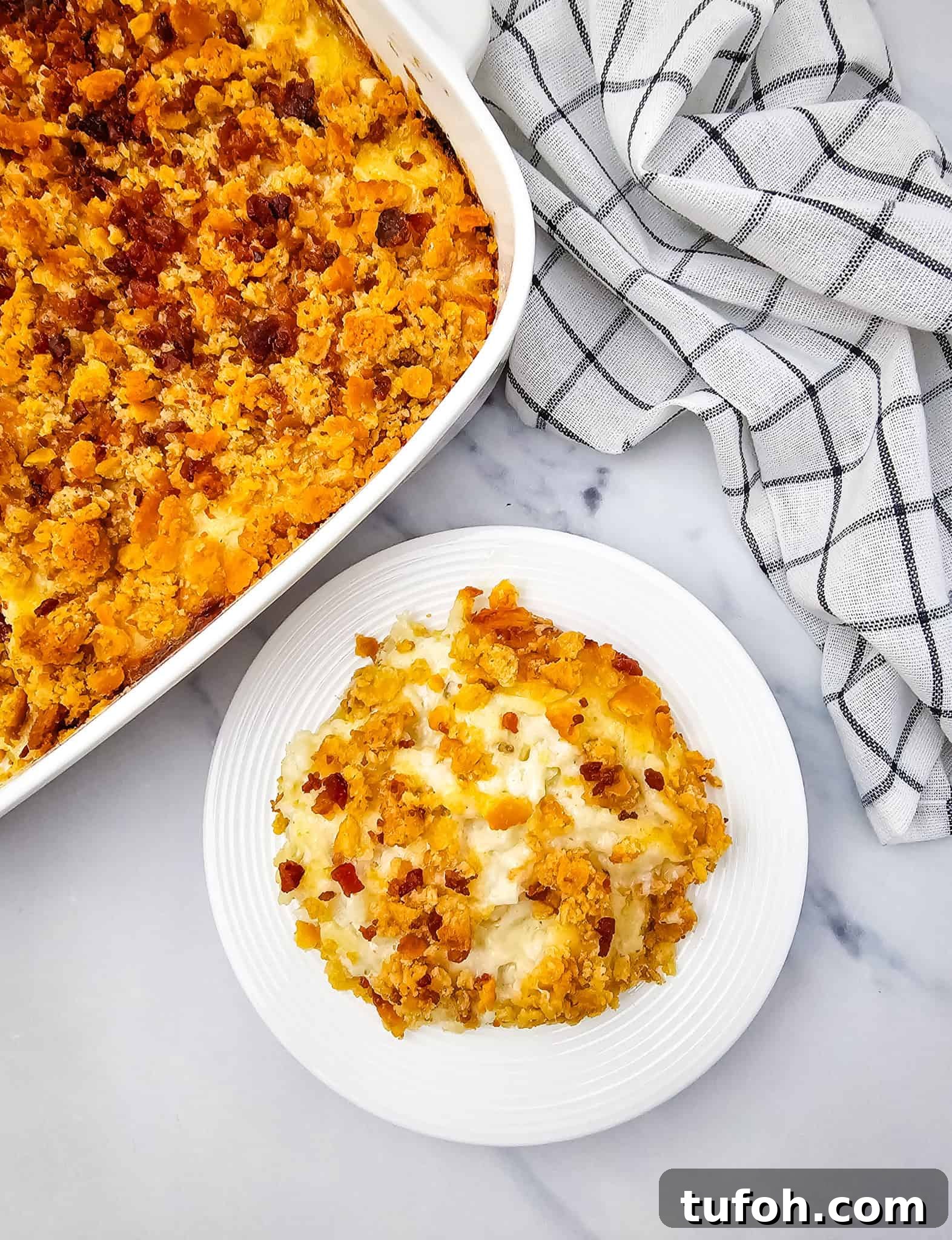 Creamy Cheesy Potato Casserole 3 A serving of cheese potato casserole with a crispy topping sits on a white plate next to the casserole dish, showcasing layers of gooey cheese perfection.