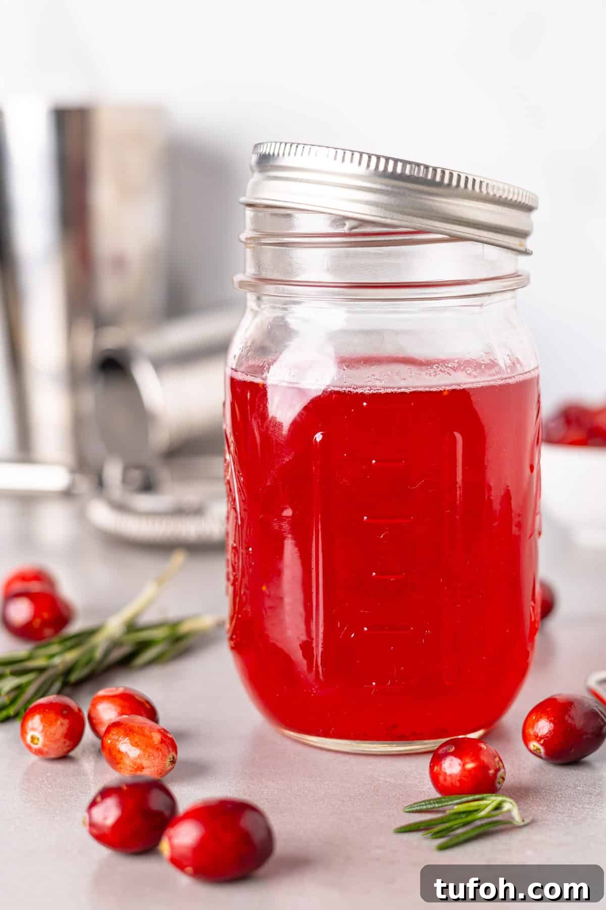 A close up shot of a jar with cranberry simple syrup on a gray table with fresh cranberries on the table.