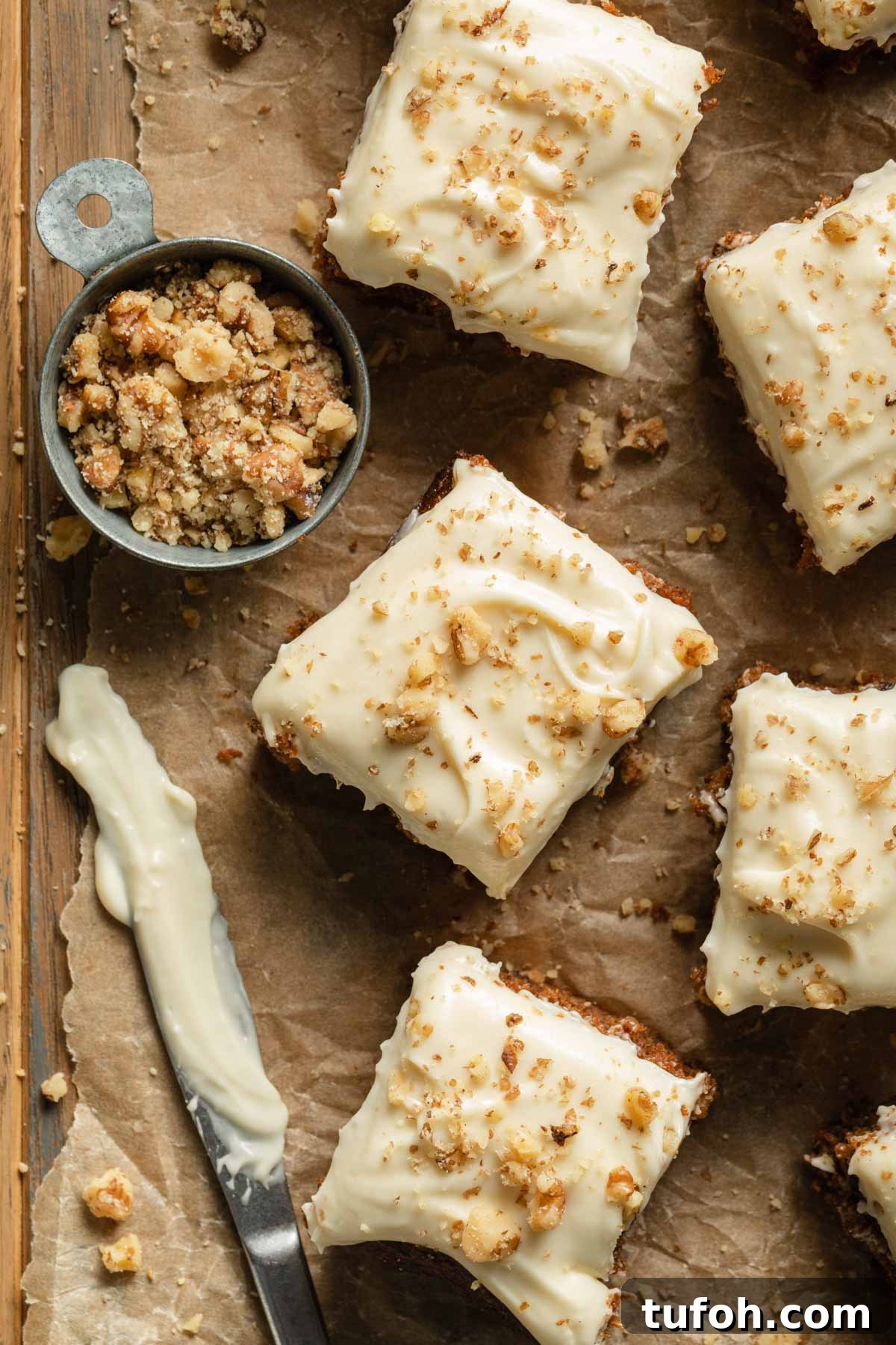 Overhead view of pieces of carrot cake arranged on a sheet of crumpled brown parchment paper next to a cup of chopped walnuts.