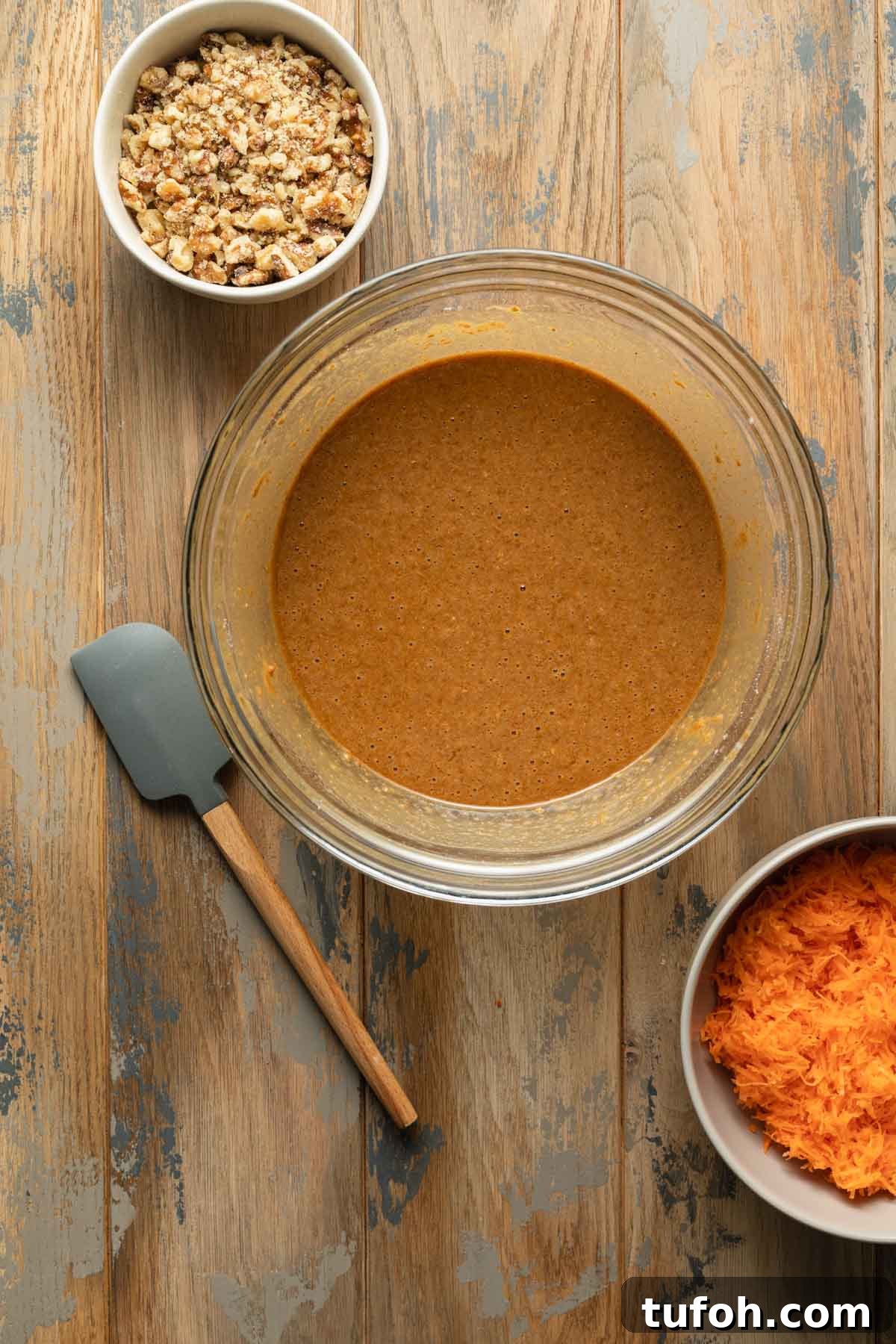 Carrot cake batter in a glass bowl next to bowls of shredded carrots and chopped walnuts.