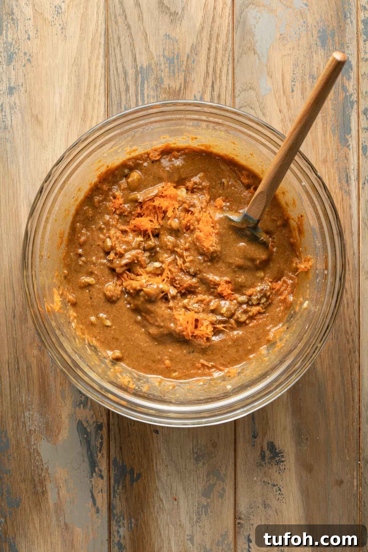 Carrot cake batter in a glass bowl with carrots and walnuts mixed in with a spatula.
