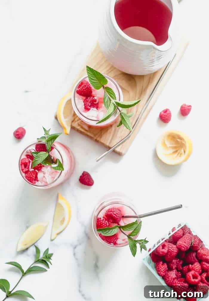 Overhead view of three glasses of raspberry lemonade on a white counter, surrounded by lemon slices and fresh raspberries