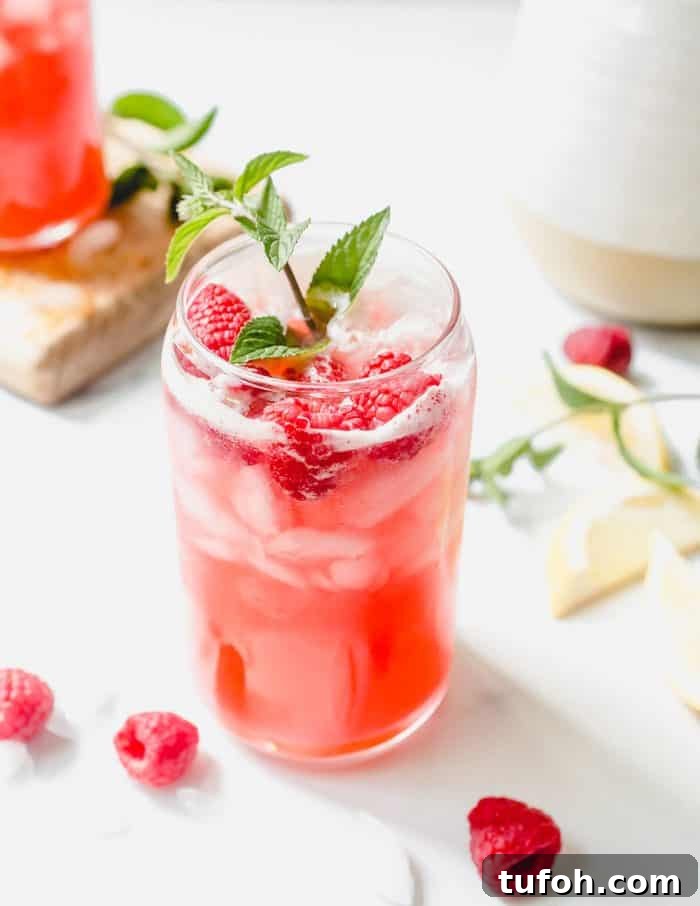Glass of raspberry lemonade garnished with a sprig of mint on a white counter, with raspberries scattered on the countertop