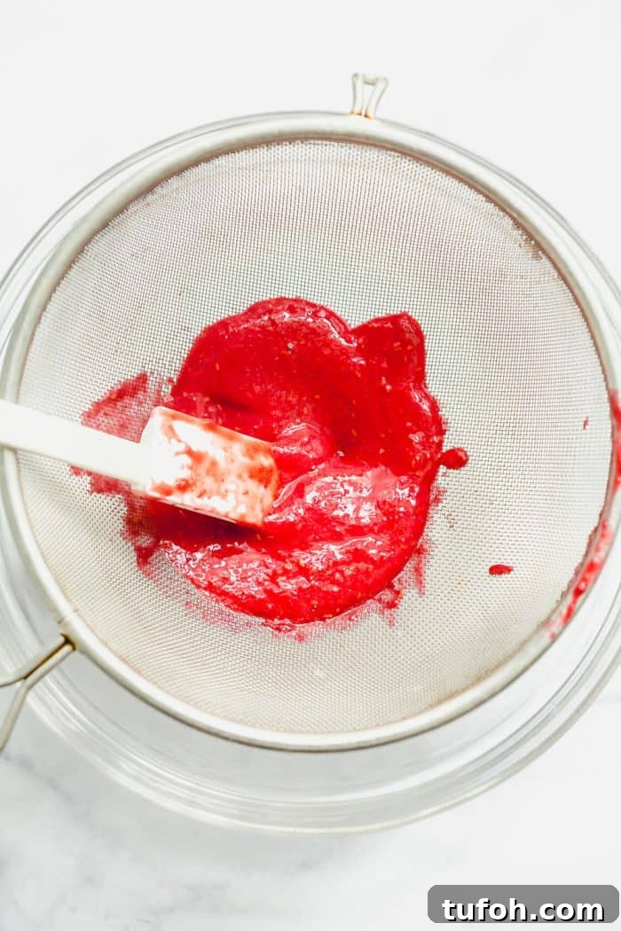 Spatula pushing raspberry puree through a fine mesh sieve set over a glass bowl on a white surface