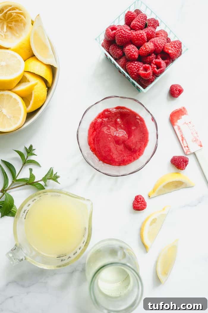 Overhead view of a bowl of raspberry puree and a measuring cup of lemon juice on a white counter, next to containers of fresh raspberries and halved lemons