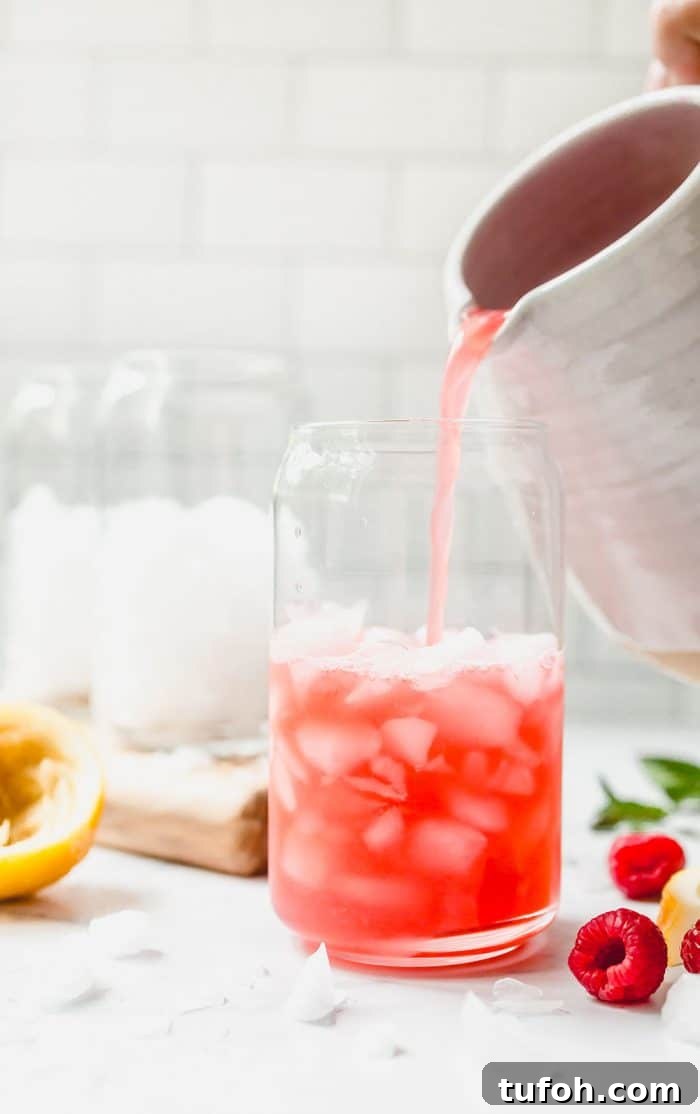 White pitcher pouring raspberry lemonade into a glass. Additional glasses with ice are in the background