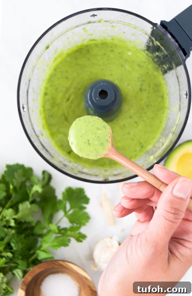 Creamy Avocado Lime Dressing Recipe for Salads & Bowls 5 A person holds a spoon of Lime Avocado Dressing above a food processor