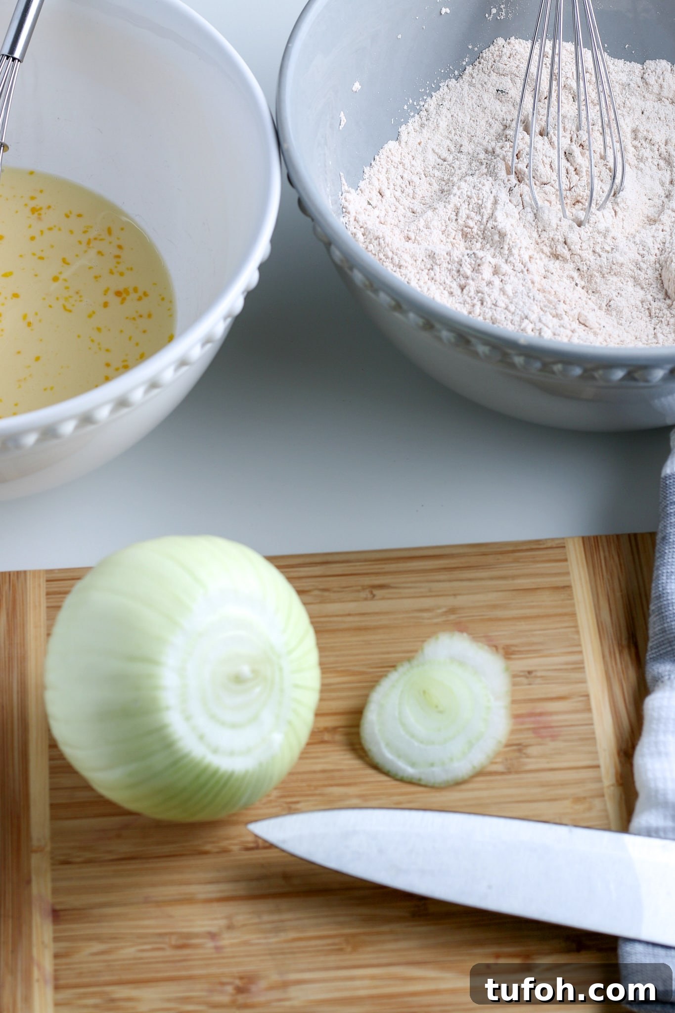 Crispy Air Fryer Bloomin' Onion Recipe for Game Day 4 a large onion on a cutting board with the top cut off, with egg wash and a flour mixture in the background