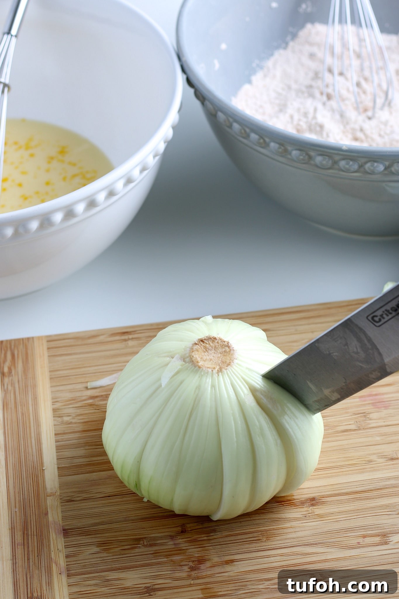 Crispy Air Fryer Bloomin' Onion Recipe for Game Day 6 raw onion being cut into a bloom on a cutting board with two bowls in the background.
