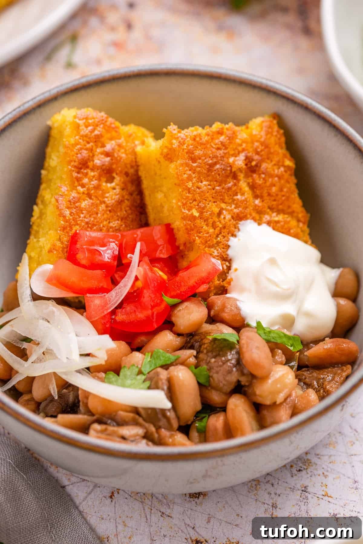 Close up of beans and cornbread in a white bowl with sour cream and onions on top.