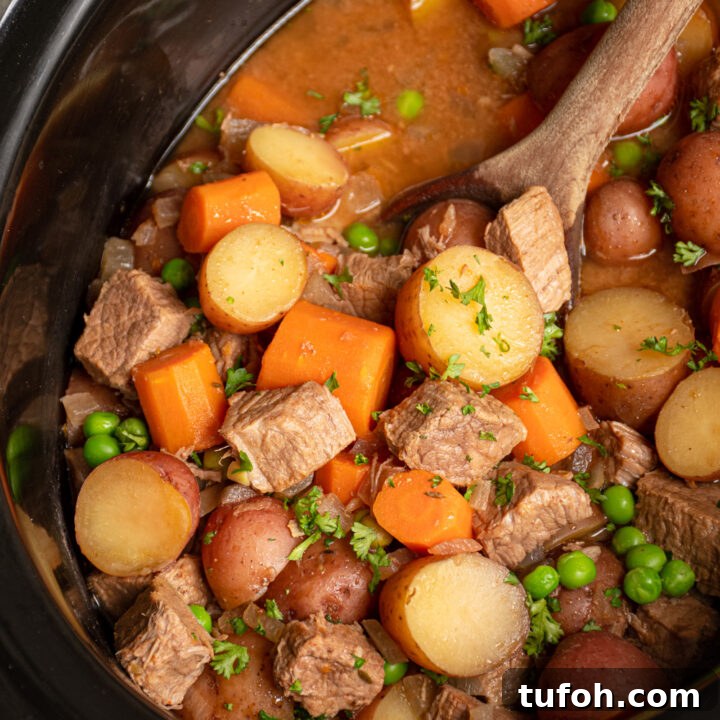Black crockpot filled with beef stew with big chunks of vegetables and stew meat and a wood spoon scooping them.