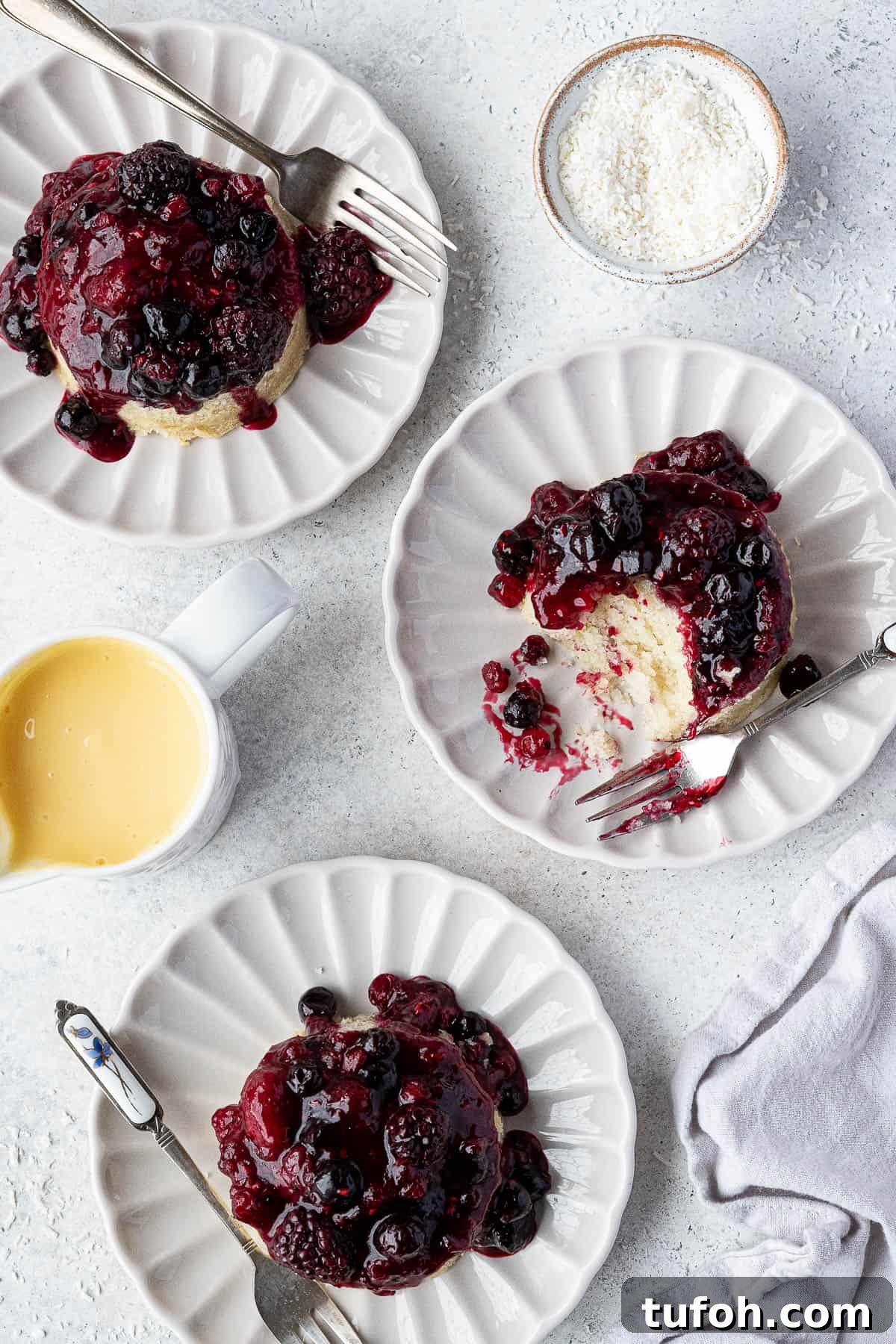 Vegan Berry & Coconut Steamed Puddings Recipe 10 Three steamed berry and coconut puddings on white plates with a jug of custard and a bowl of coconut.