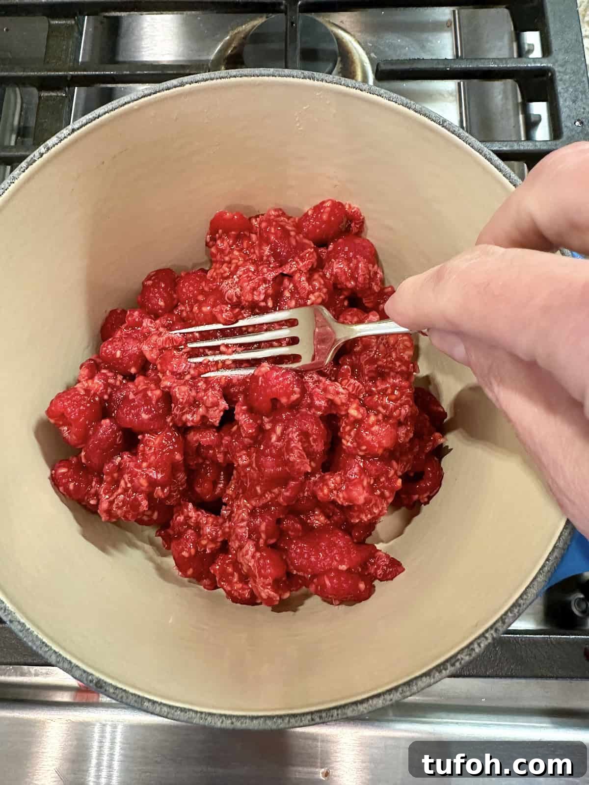 No-Cook Raspberry Jam Without Pectin or Canning 5 A fork mashing fresh raspberries in a pan on the stove.