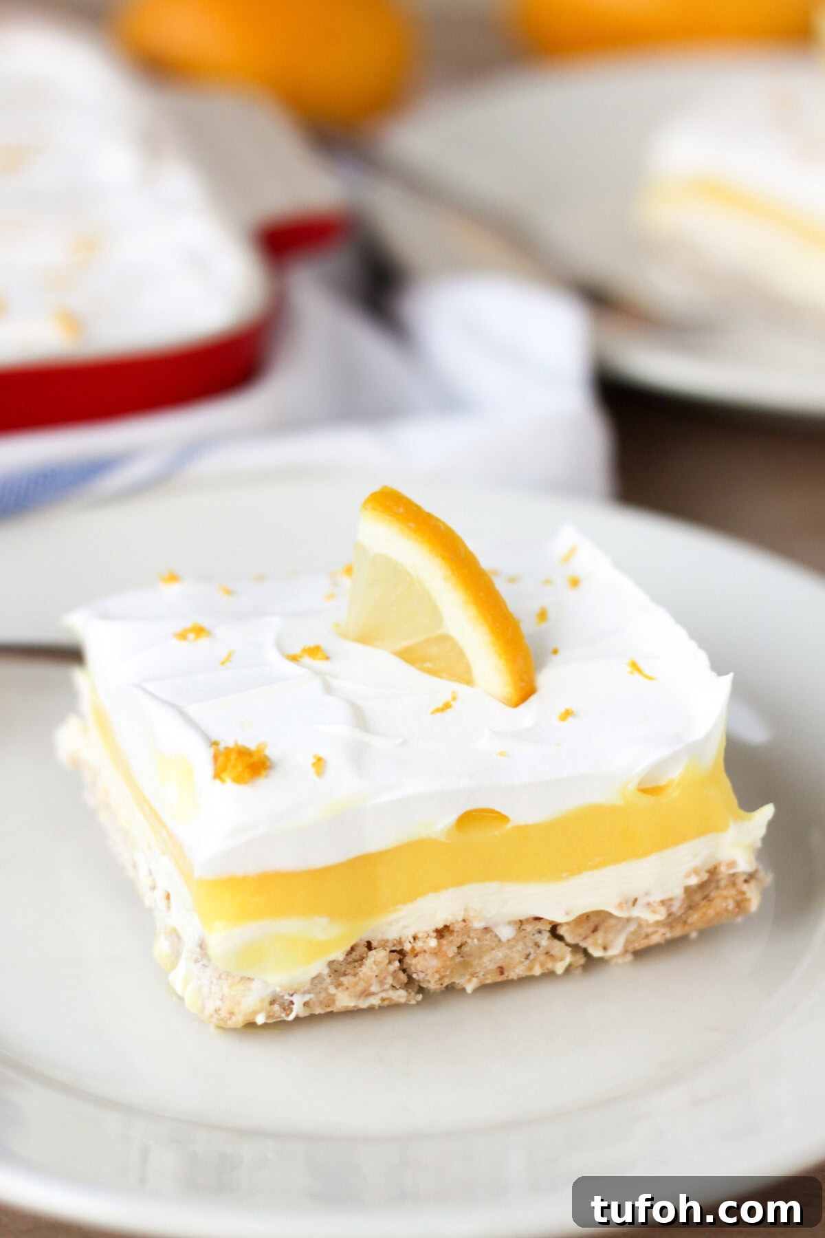 Lemon Lush. It's light and refreshing with 4 layers consisting of bottom pecan crust, lemon cream cheese, lemon pudding and topped with a whipped layer garnished with lemon zest. #lemonlush #lemondessert #lemonlasagna #nobake #onepandesserts #potluckdesserts #creamcheesedesserts #puddingdesserts Piece of Lemon Lush on a plate