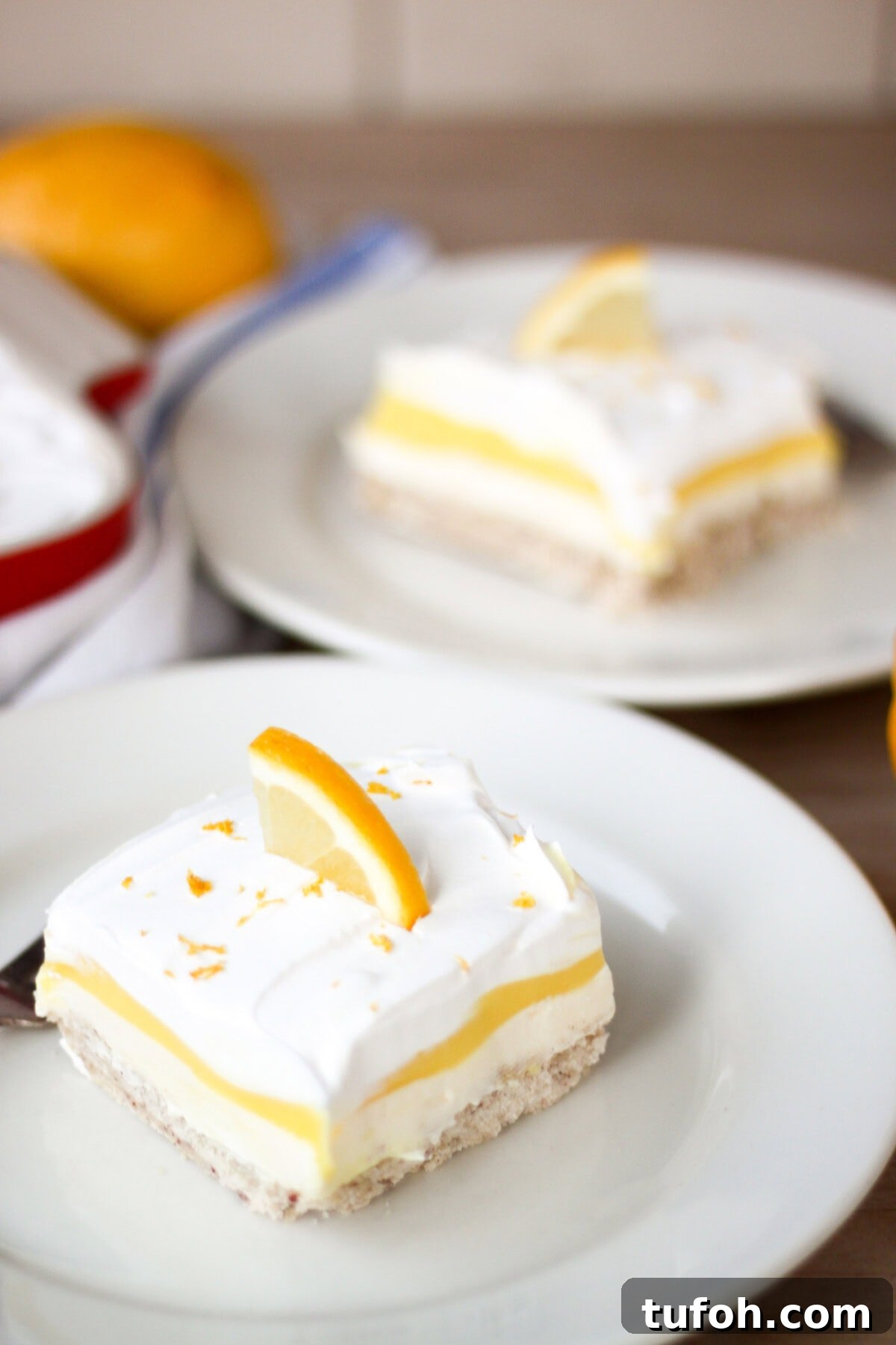 Lemon Lush. It's light and refreshing with 4 layers consisting of bottom pecan crust, lemon cream cheese, lemon pudding and topped with a whipped layer garnished with lemon zest. #lemonlush #lemondessert #lemonlasagna #nobake #onepandesserts #potluckdesserts #creamcheesedesserts #puddingdesserts Two pieces of Lemon Lush on plates