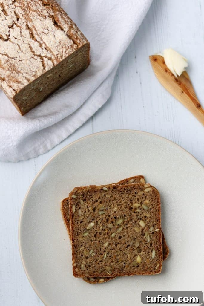 Sourdough Danish Rugbrød Rye Loaf Recipe 2 Rye bread slices on a plate next to a loaf of rye bread and a wooden knife with butter