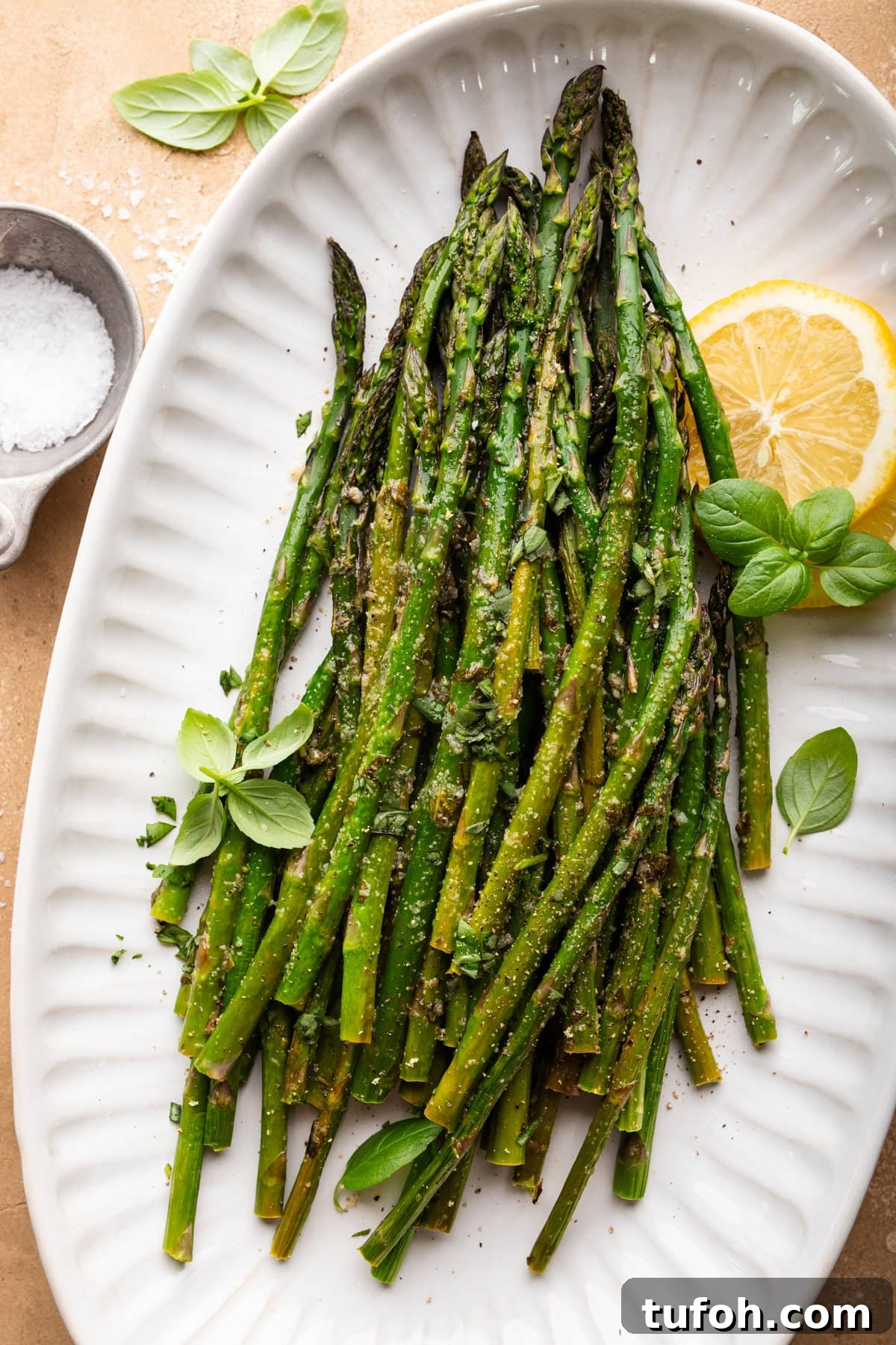 A large white plate with roasted asparagus on it. The asparagus is topped with fresh basil leaves and lemon slices. Arranged next to the plate is a small dish of salt and a fresh basil leaf.