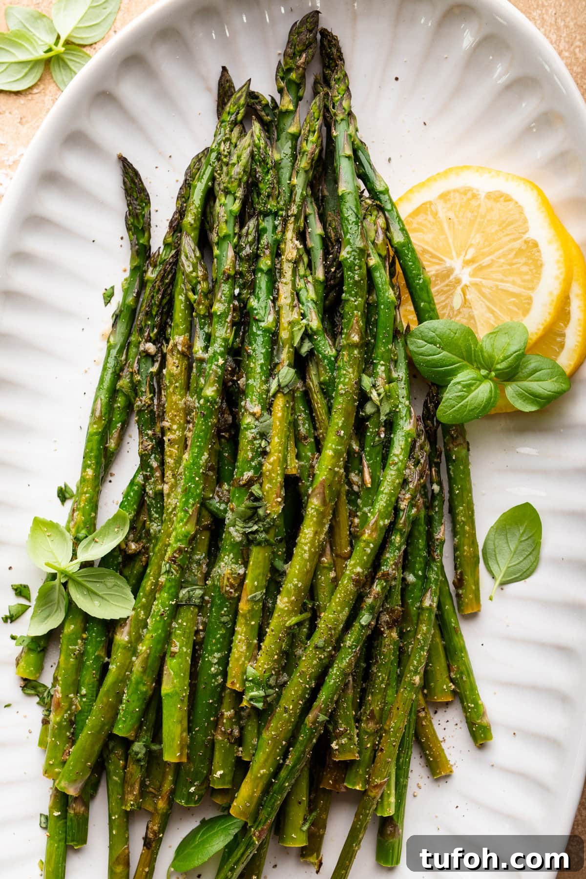 Roasted asparagus arranged on a white plate with fresh herbs and lemon slices arranged around the asparagus.