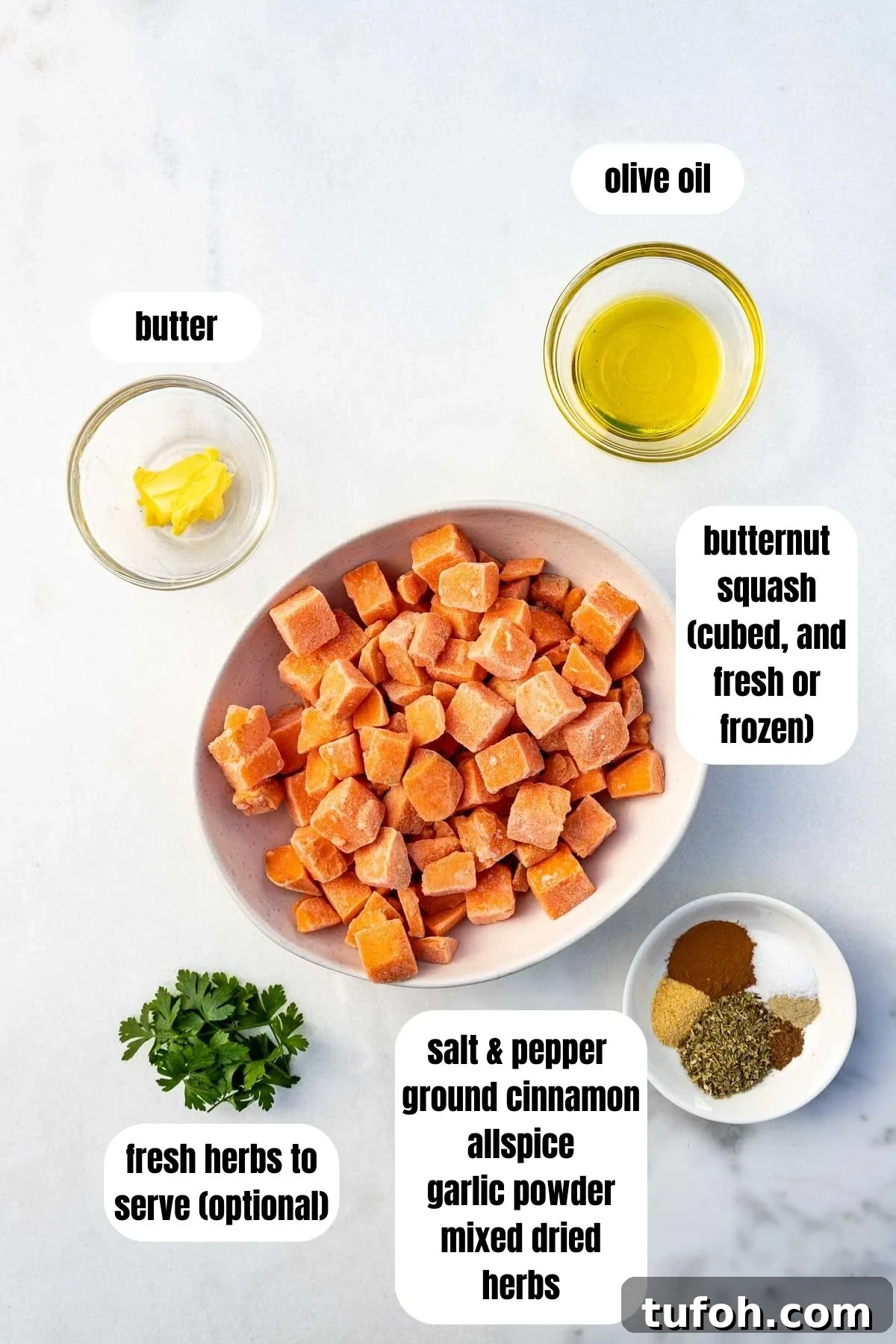 Overhead of all the ingredients needed to sautee butternut squash including the cubed frozen butternut squash, olive oil, a little butter, salt and pepper, ground cinnamon, allspice, garlic powder, mixed dried herbs and fresh herbs to serve, all on a marble background.