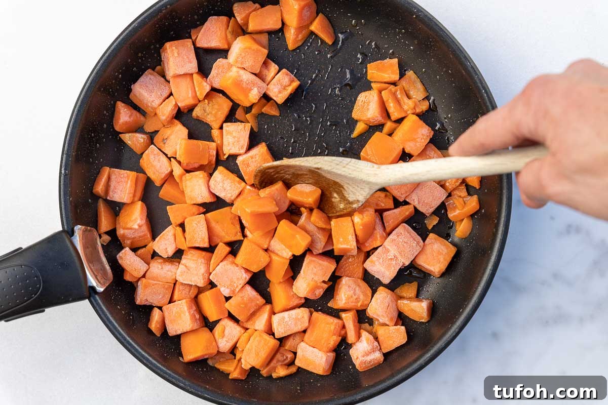 Overhead of someone's hand stirring a frying pan of cubed frozen butternut squash with a wooden spoon, all on a marble background.