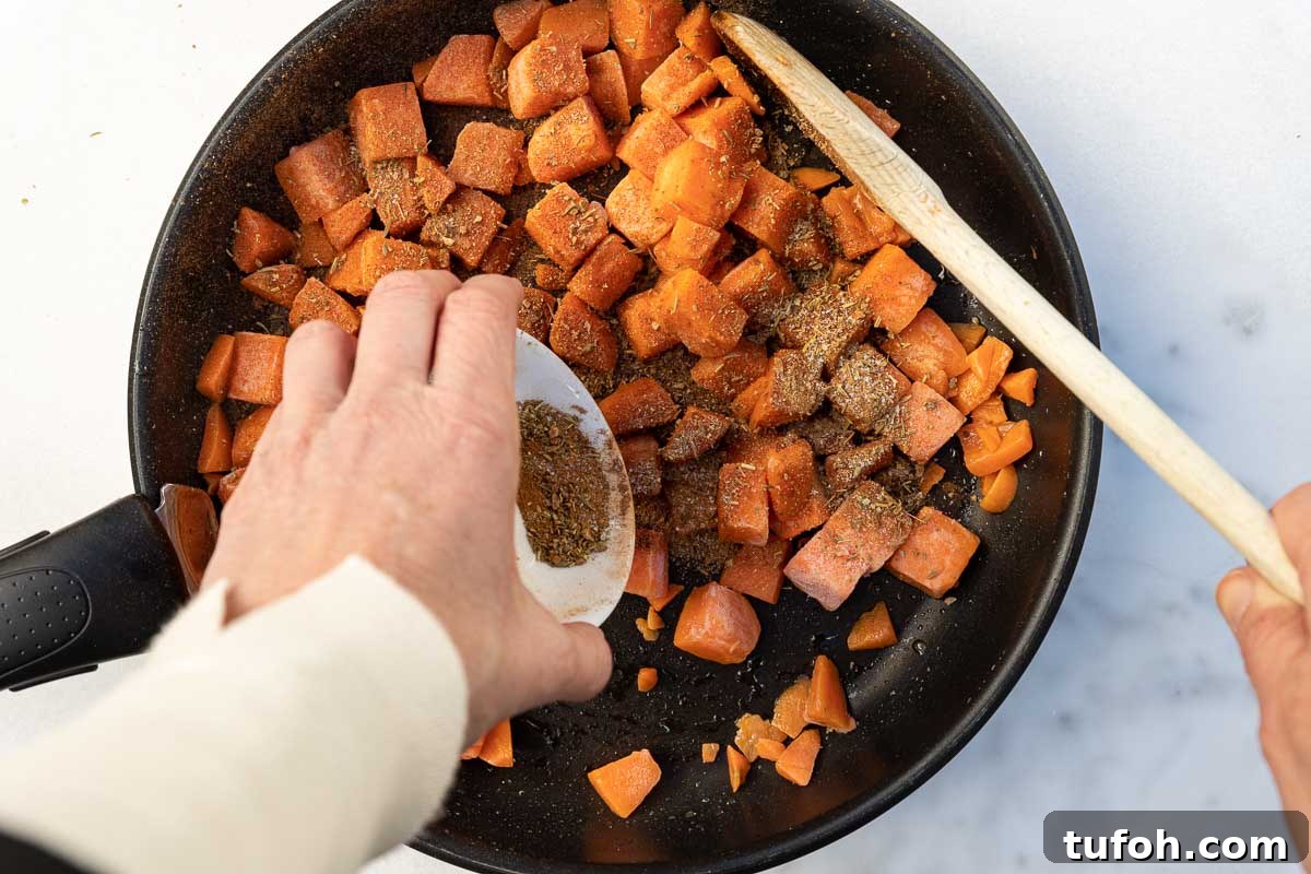 Someone's hand adding a small white pot of mixed herbs and spices to a frying pan of cubed butternut squash with a wooden spoon and on a marble background.