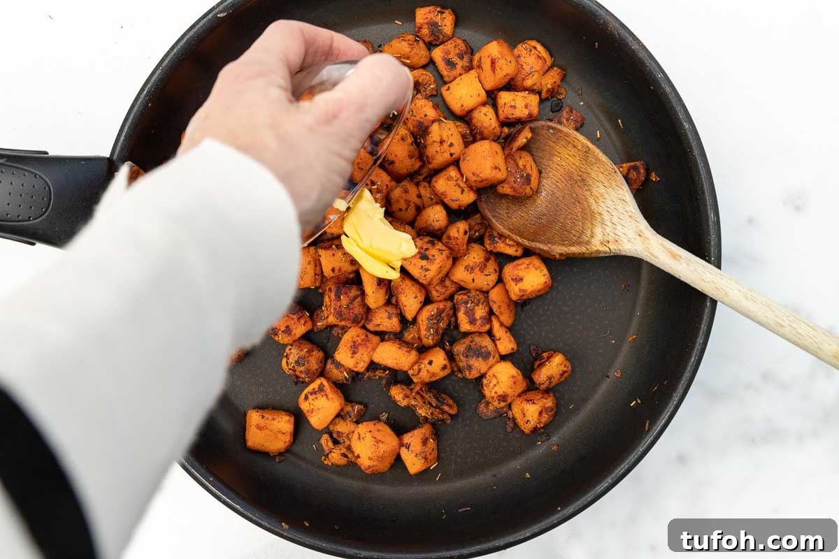 Overhead of someone's arm and hand shown adding a little butter to a pan of sauteed cubed butternut squash in a frying pan with a wooden spoon.