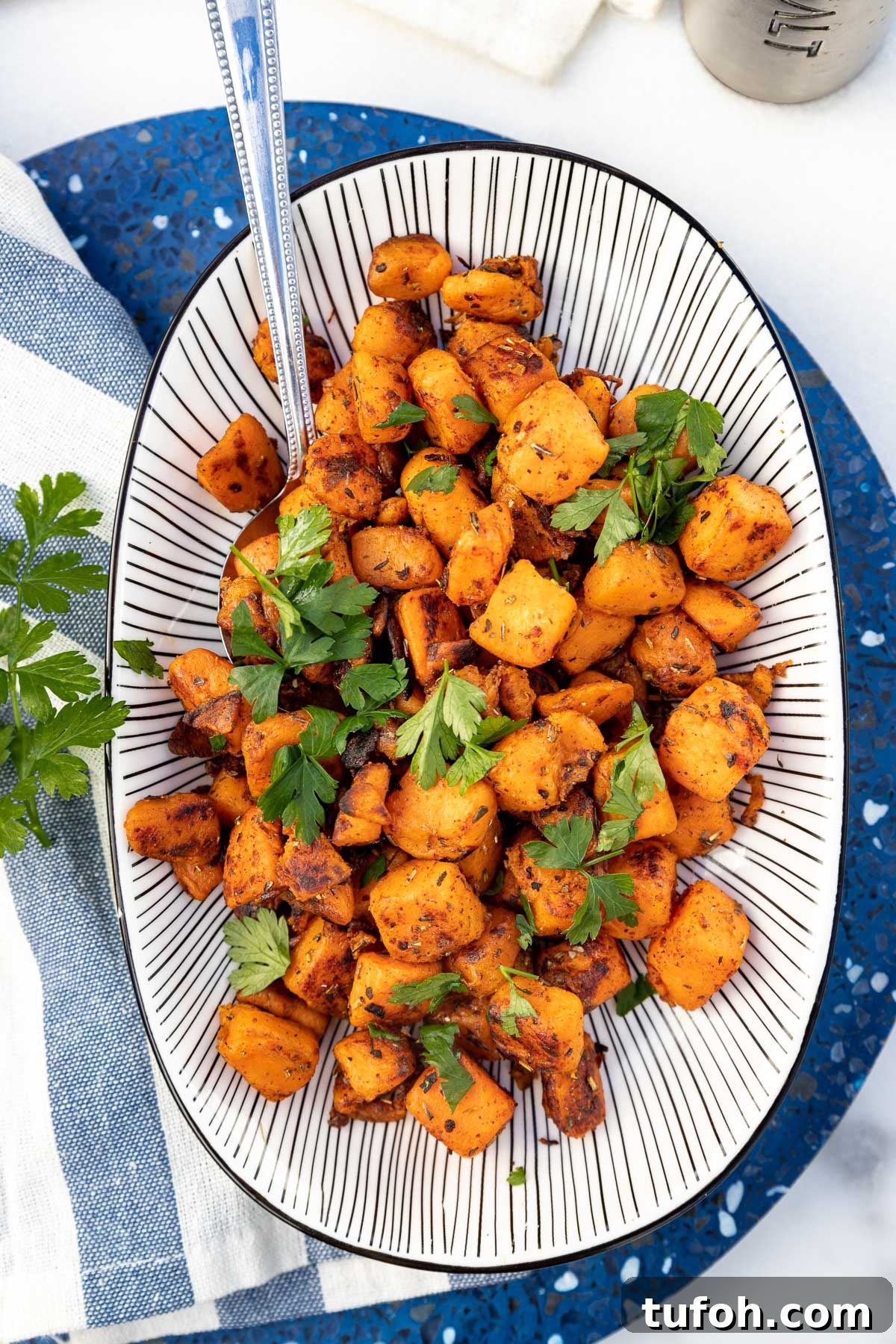 An overhead view of a black and white plate of cubed sauteed butternut squash cubes with fresh parsley on top, all on a blue marble background with blue and white tea towel on the left.
