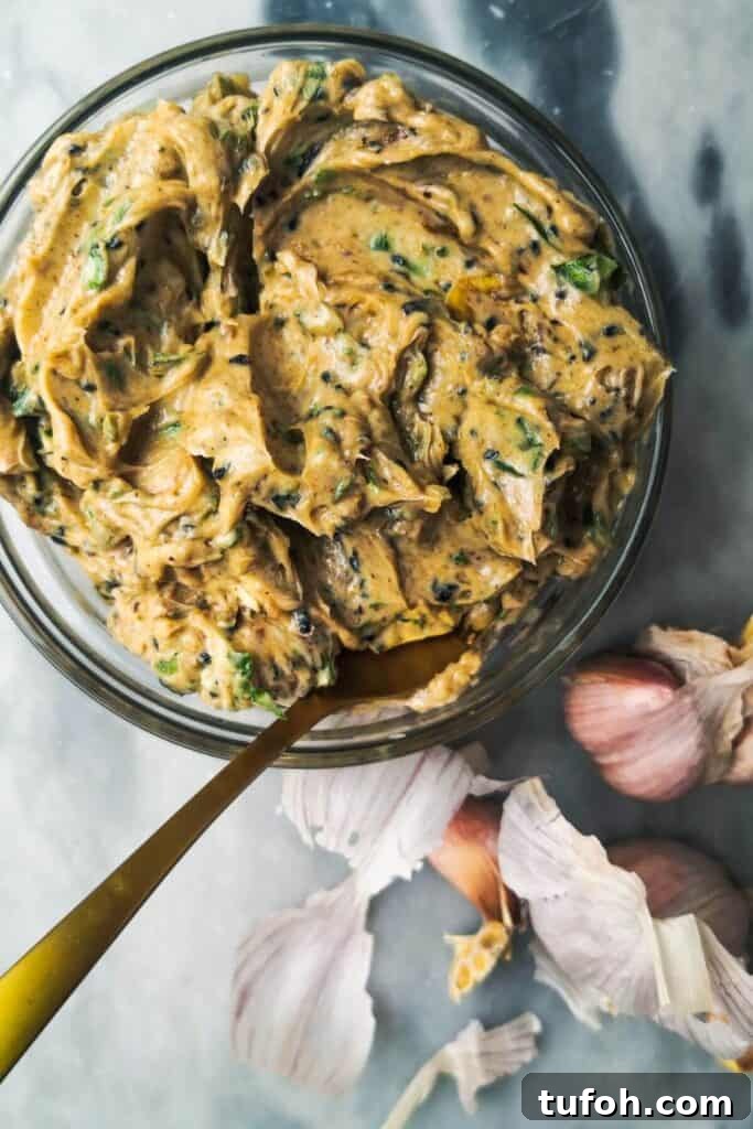 Roasted garlic butter in a small glass bowl with a gold spoon inside, with garlic cloves in the background.