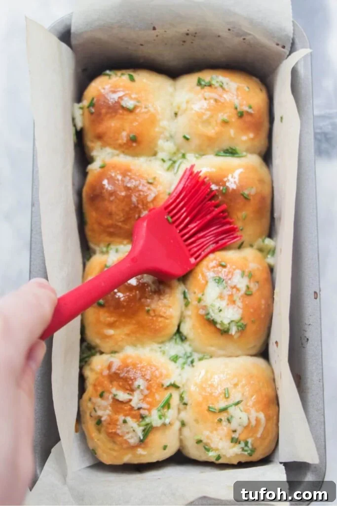 Garlic butter being brushed on 8 golden dinner rolls in a baking paper lined tin with a red brush.