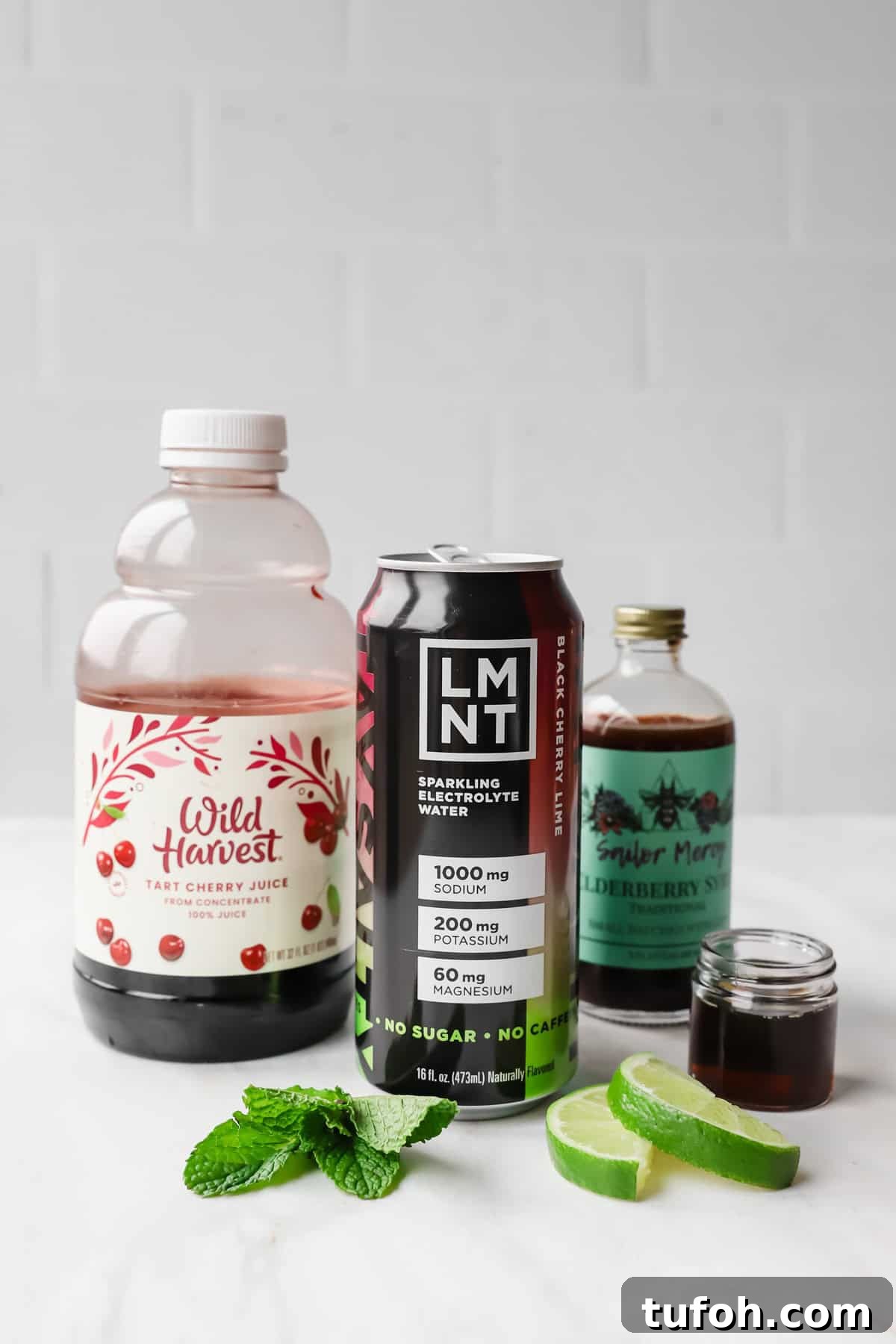 Relaxing Sleepy Girl Mocktail Recipe — Caffeine-Free Nightcap 3 A bottle of tart cherry juice, a can of LMNT electrolyte drink, elderberry syrup, mint leaves, and lime slices on a white surface.