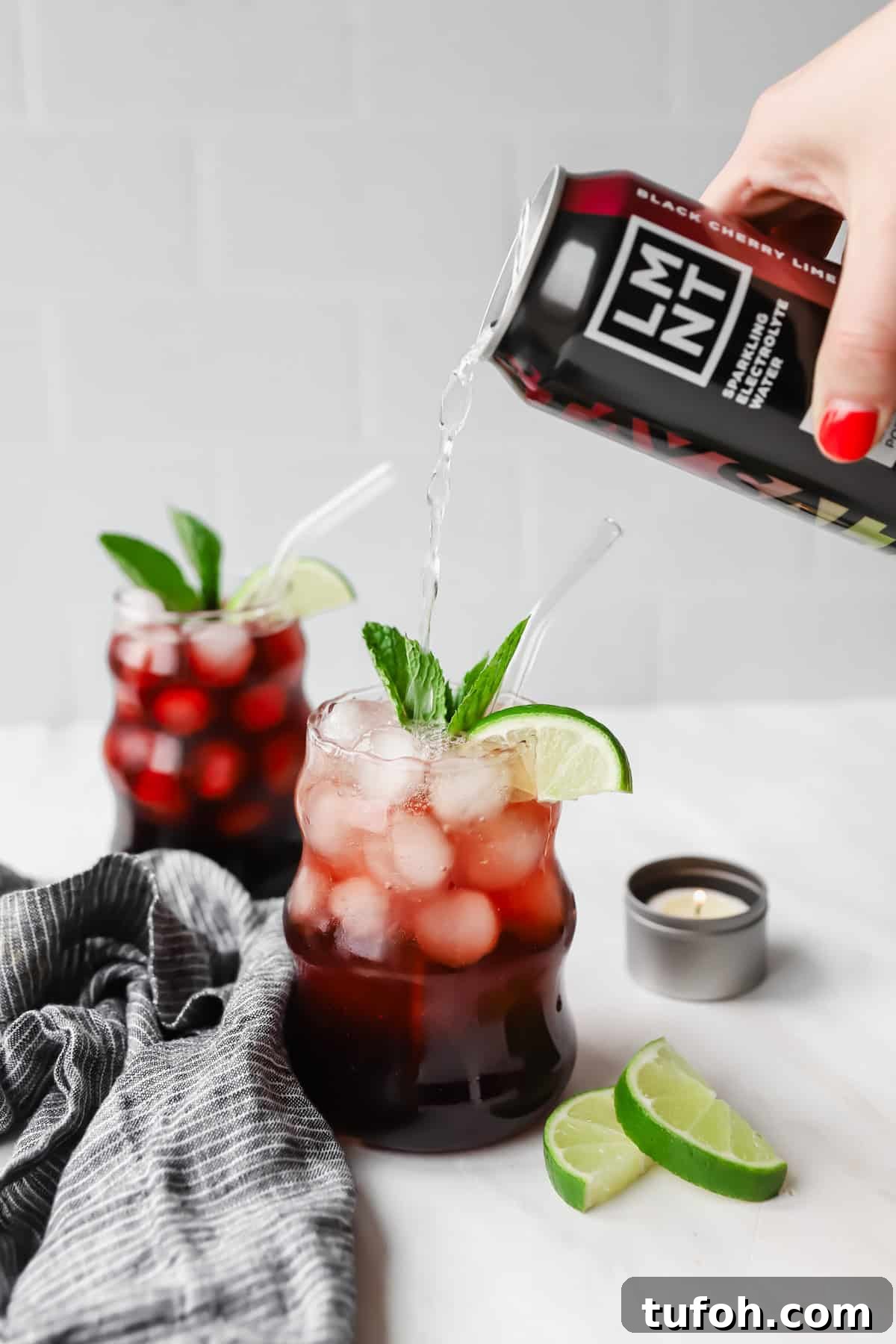 Relaxing Sleepy Girl Mocktail Recipe — Caffeine-Free Nightcap 5 A hand pours a black cherry drink into a glass filled with ice, cherries, lime slices, and mint. Another similar drink is in the background, next to lime wedges and a small candle.
