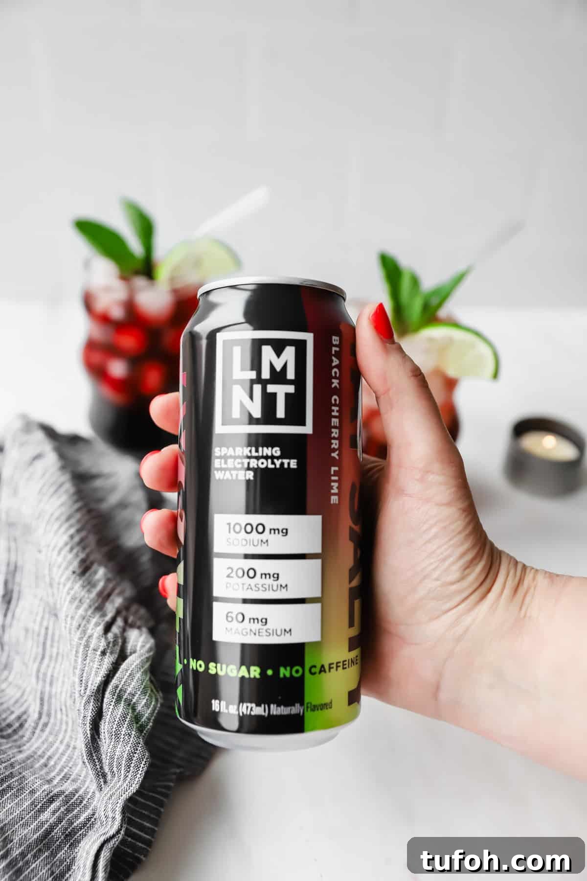 Relaxing Sleepy Girl Mocktail Recipe — Caffeine-Free Nightcap 6 Hand holding a can of LMNT black cherry lime sparkling electrolyte water. Two glasses with red drinks and lime wedges are in the background.