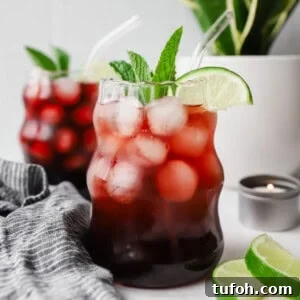 Relaxing Sleepy Girl Mocktail Recipe — Caffeine-Free Nightcap 7 Two glasses of iced berry drink with lime slices and mint, placed on a table with a striped cloth. A small candle and lime wedges are nearby, with a potted plant in the background.