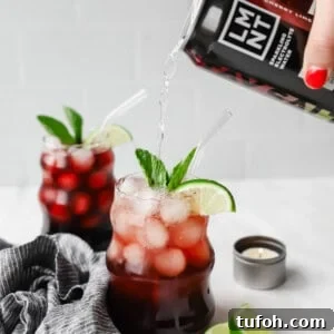 Relaxing Sleepy Girl Mocktail Recipe — Caffeine-Free Nightcap 9 A hand pours a black cherry drink into a glass filled with ice, cherries, lime slices, and mint. Another similar drink is in the background, next to lime wedges and a small candle.