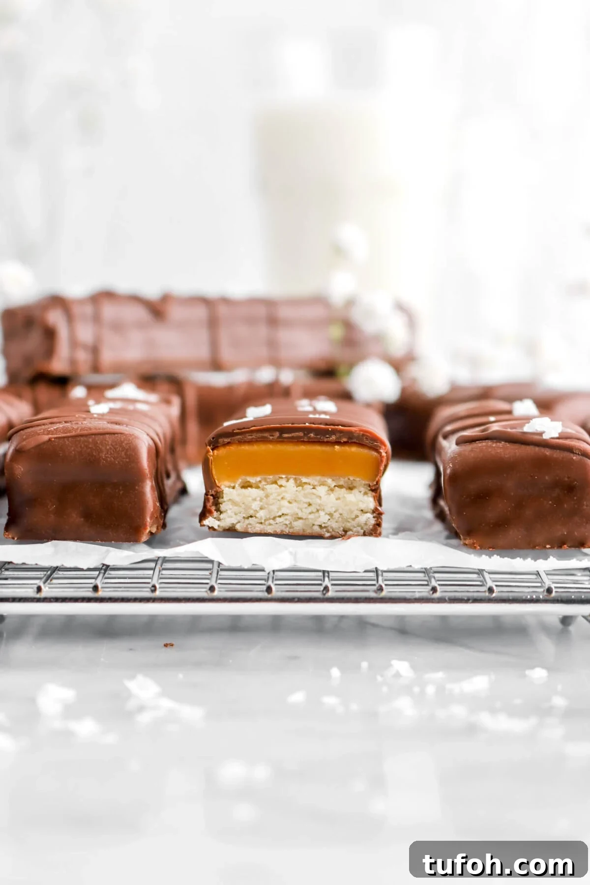 close up of cut twix bar on wire cooling rack with more bars behind, a glass of milk, and white flowers
