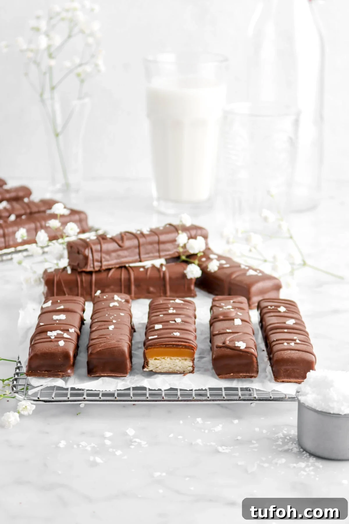 pulled back shot of twix bars with one cut open on wire cooling rack with flowers, glass of milk, and flaked salt