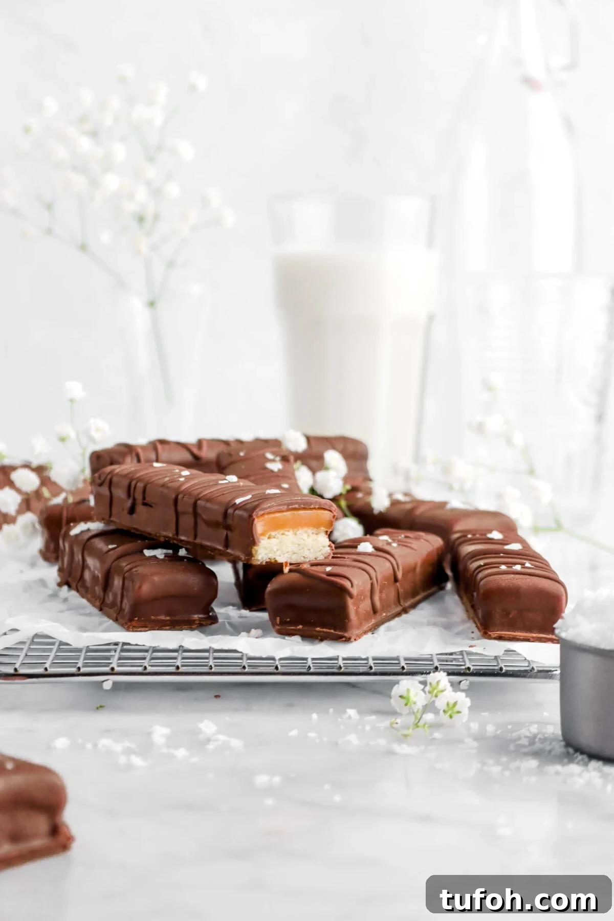 homemade twix bars on wire cooling rack on parchment paper with one cut open, flaked salt, flowers, and a glass of milk behind