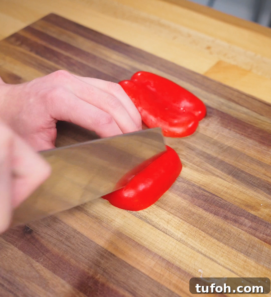 Beijing-Style Beef Stir-Fry Recipe 5 A hand using a sharp knife to slice a vibrant red bell pepper into chunks on a wooden cutting board, prepping vegetables for Beijing Beef.