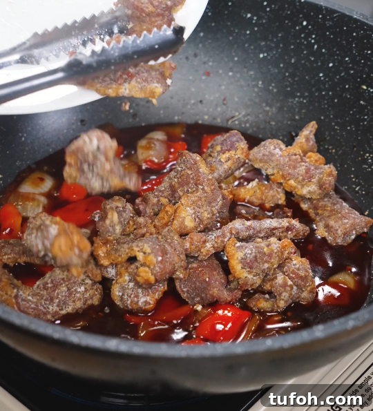 Beijing-Style Beef Stir-Fry Recipe 9 Crispy fried beef being added into a rich, bubbling Beijing Beef sauce with onions and peppers in a nonstick wok.