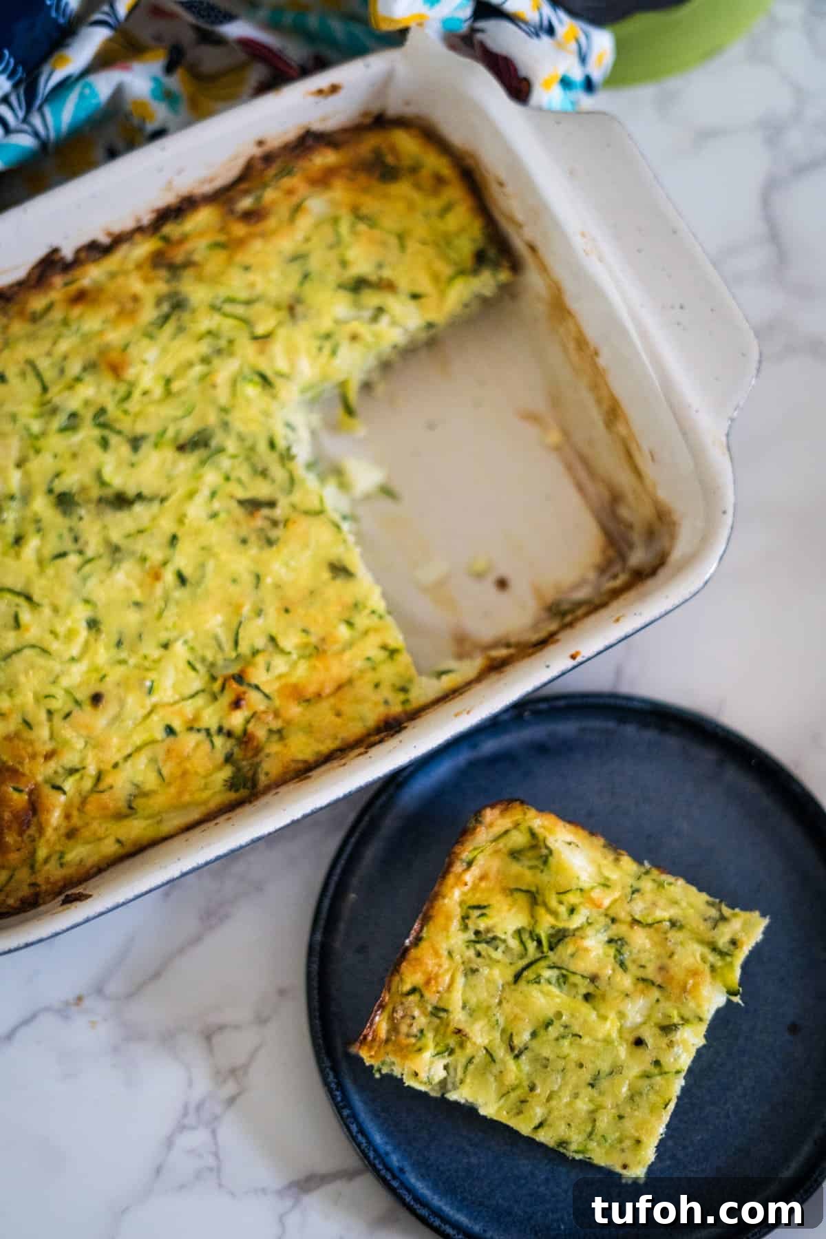 A rectangular dish of baked zucchini casserole with a piece cut out and placed on a black plate beside it. The casserole is light yellow-green with a golden-brown top.