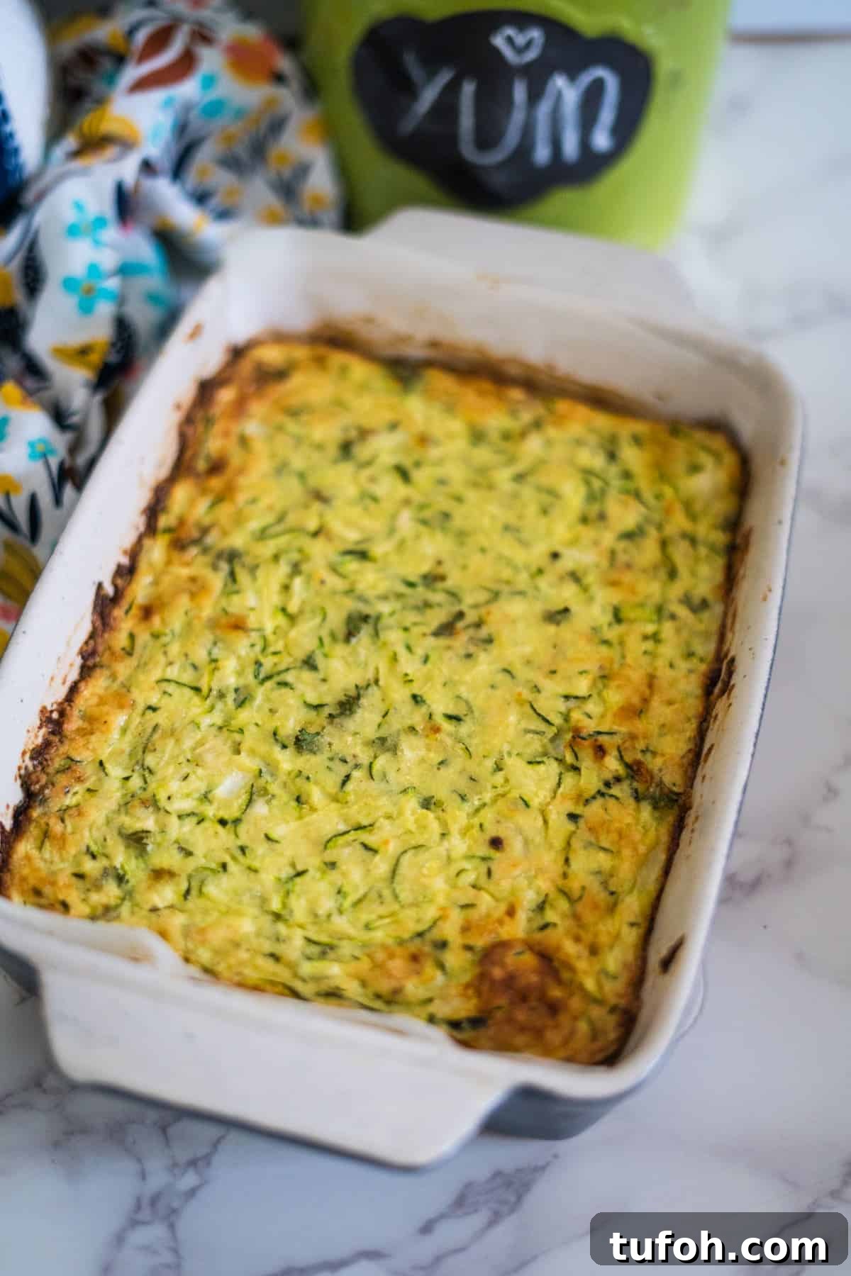 A baked casserole dish filled with a golden-brown keto zucchini slice is placed on a marble countertop. A chalkboard label in the background reads "YUM".