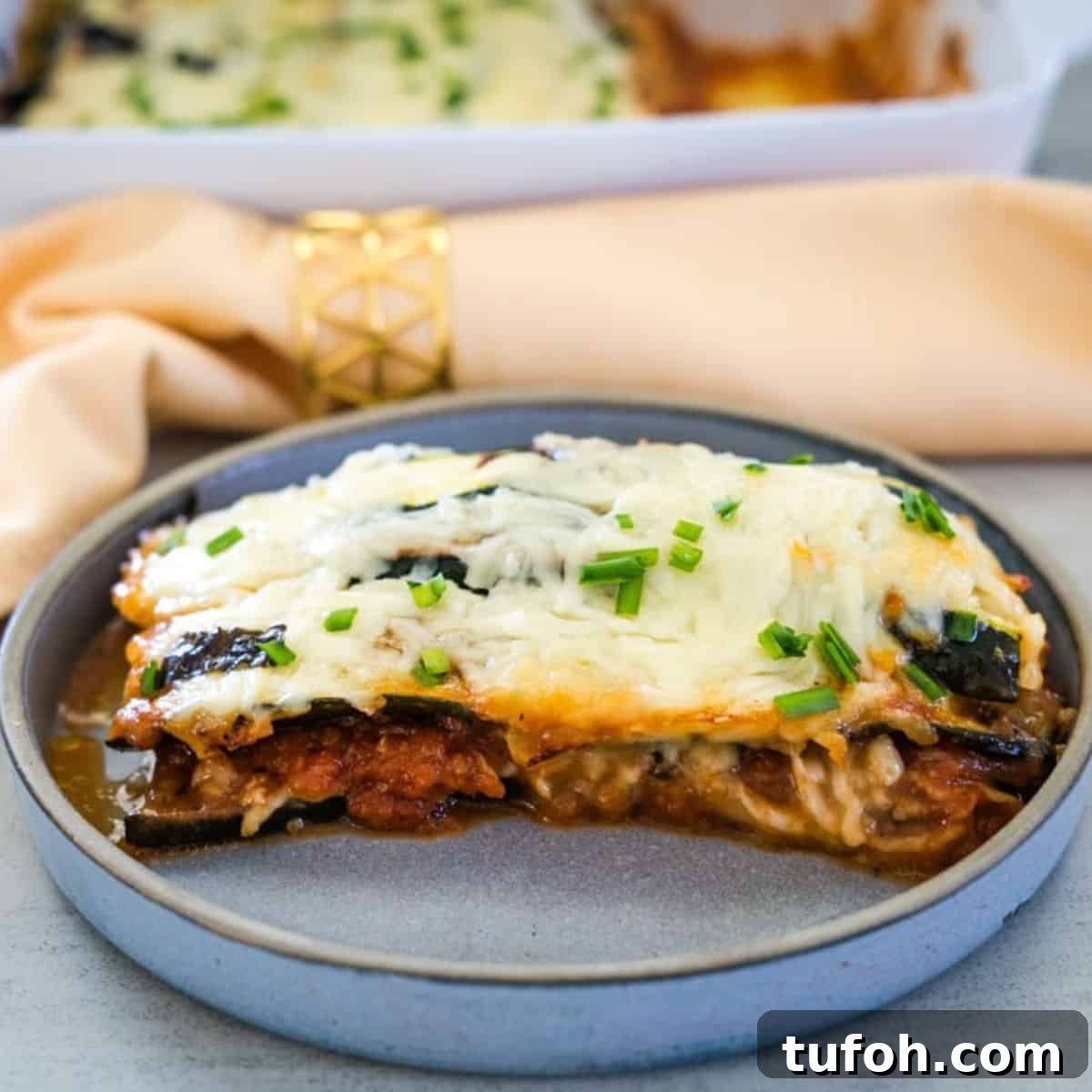 A slice of baked lasagna topped with melted cheese and chopped chives served on a grey plate, with a golden napkin ring and baking dish in the background.