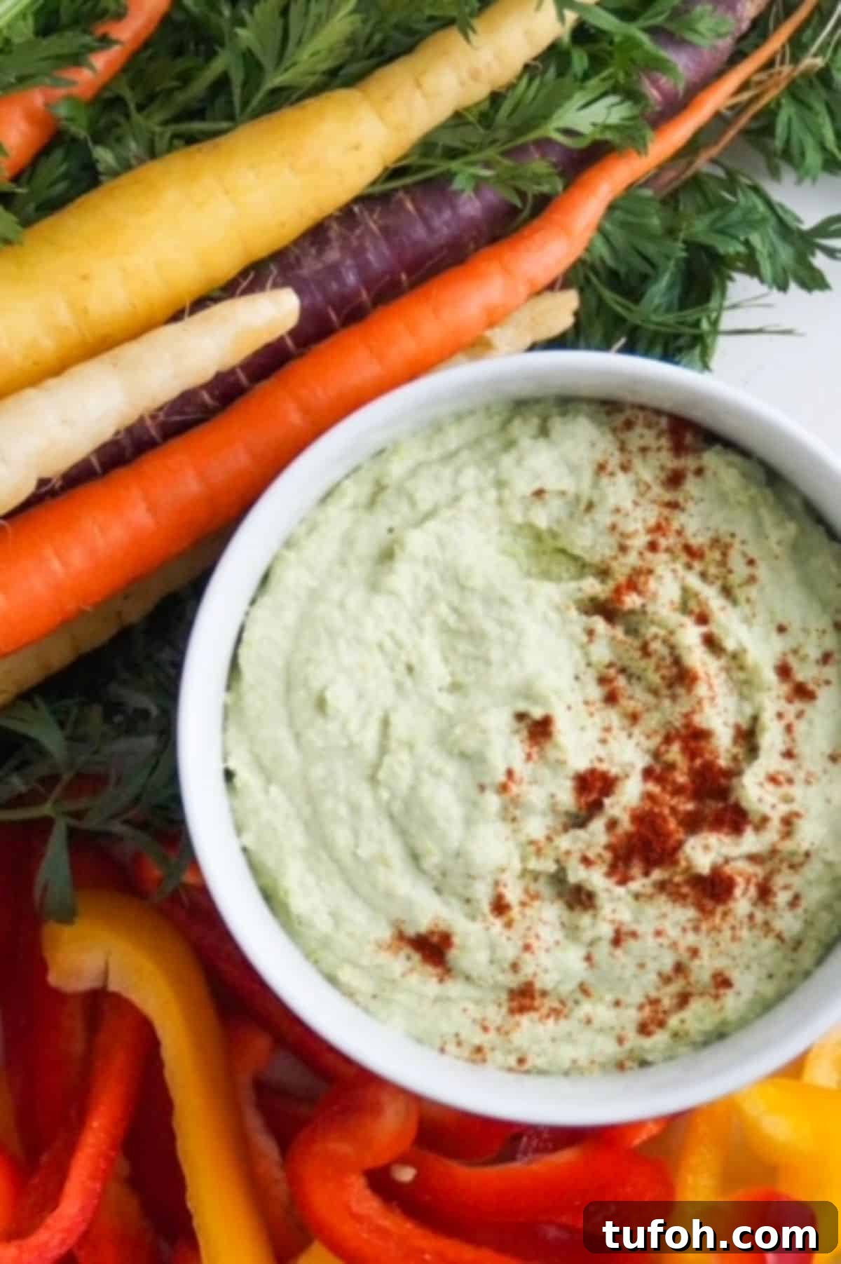 Edamame Hummus Recipe: Creamy, Protein-Packed Dip 2 Edamame hummus topped in a white bowl and topped with paprika surrounded by tri-colored carrots and sliced red and yellow bell peppers.