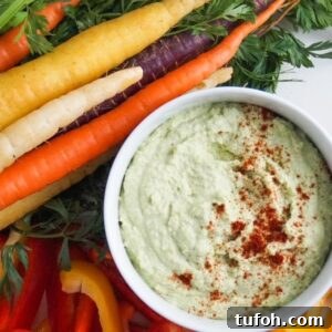 Edamame hummus in a white serving bowl and topped with paprika surrounded by tri-colored carrots and slices of bell pepper.
