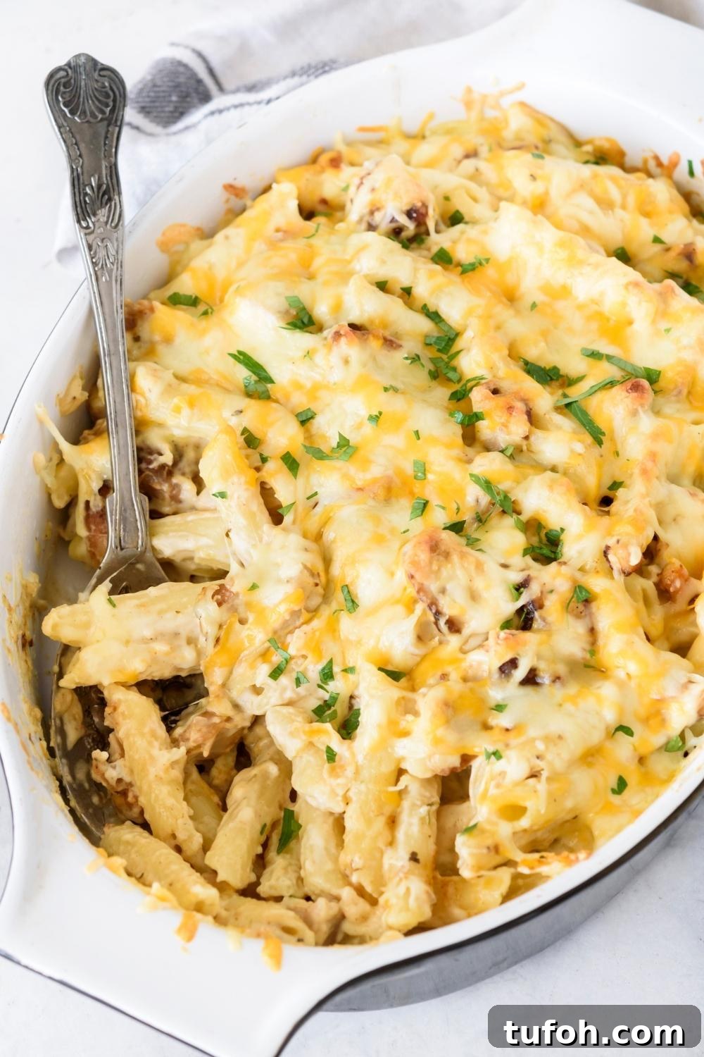 A casserole dish with chicken alfredo bake garnished with parsley. A spoon is sitting in the dish ready to serve.