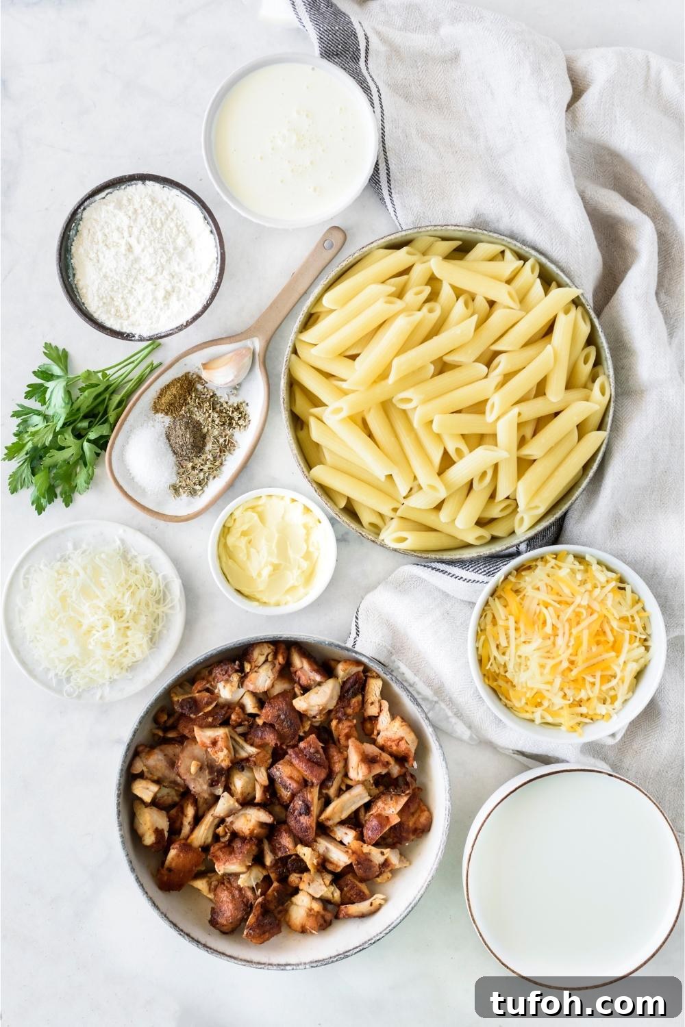 A countertop with multiple bowls containing cooked pasta, seasonings, chicken, cheese, flour, and heavy cream.