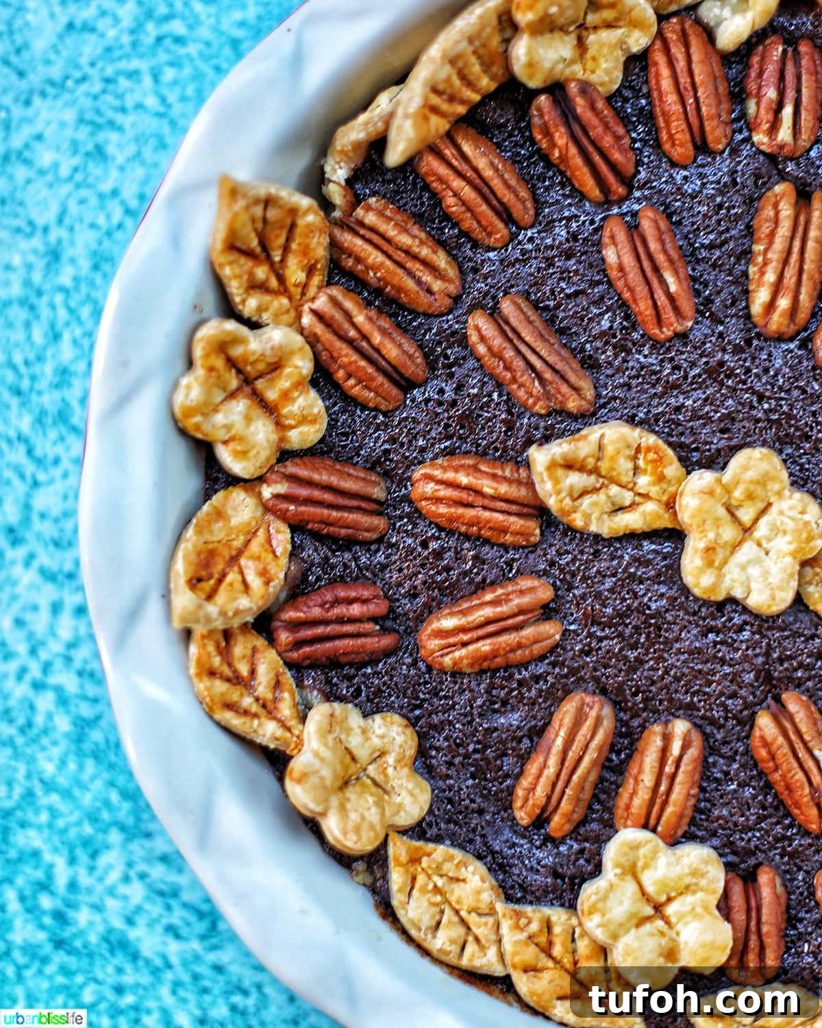 Decadent Chocolate Bourbon Pecan Pie Recipe 2 left half of Chocolate Bourbon Pecan Pie with pie crust cutouts in leaf and flower shapes with pecan halves as pie decoration in a white pie pan on blue table.