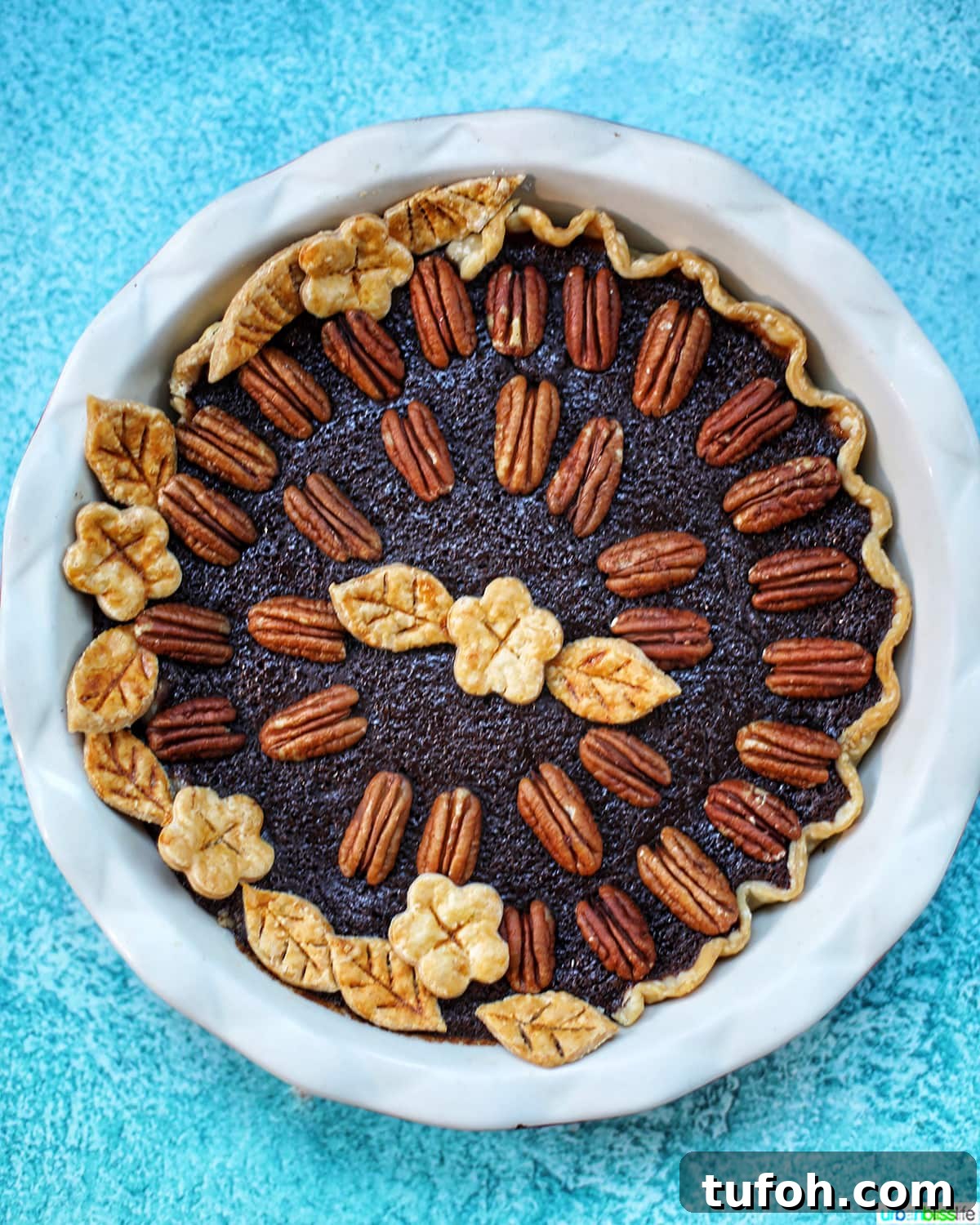 Decadent Chocolate Bourbon Pecan Pie Recipe 3 Chocolate Bourbon Pecan Pie with pie crust cutouts in leaf and flower shapes with pecan halves as pie decoration in a white pie pan on blue table.