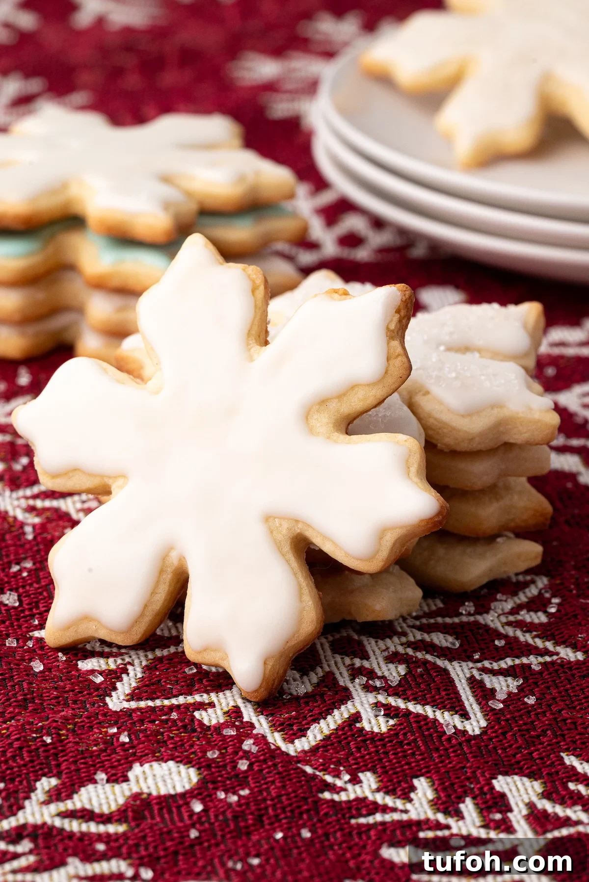 Soft Cut-Out Sugar Cookies Recipe for Tender, Perfect Shapes 11 Stack of frosted soft no-spread sugar cookies, cut into snowflake shapes on a red tablecloth.